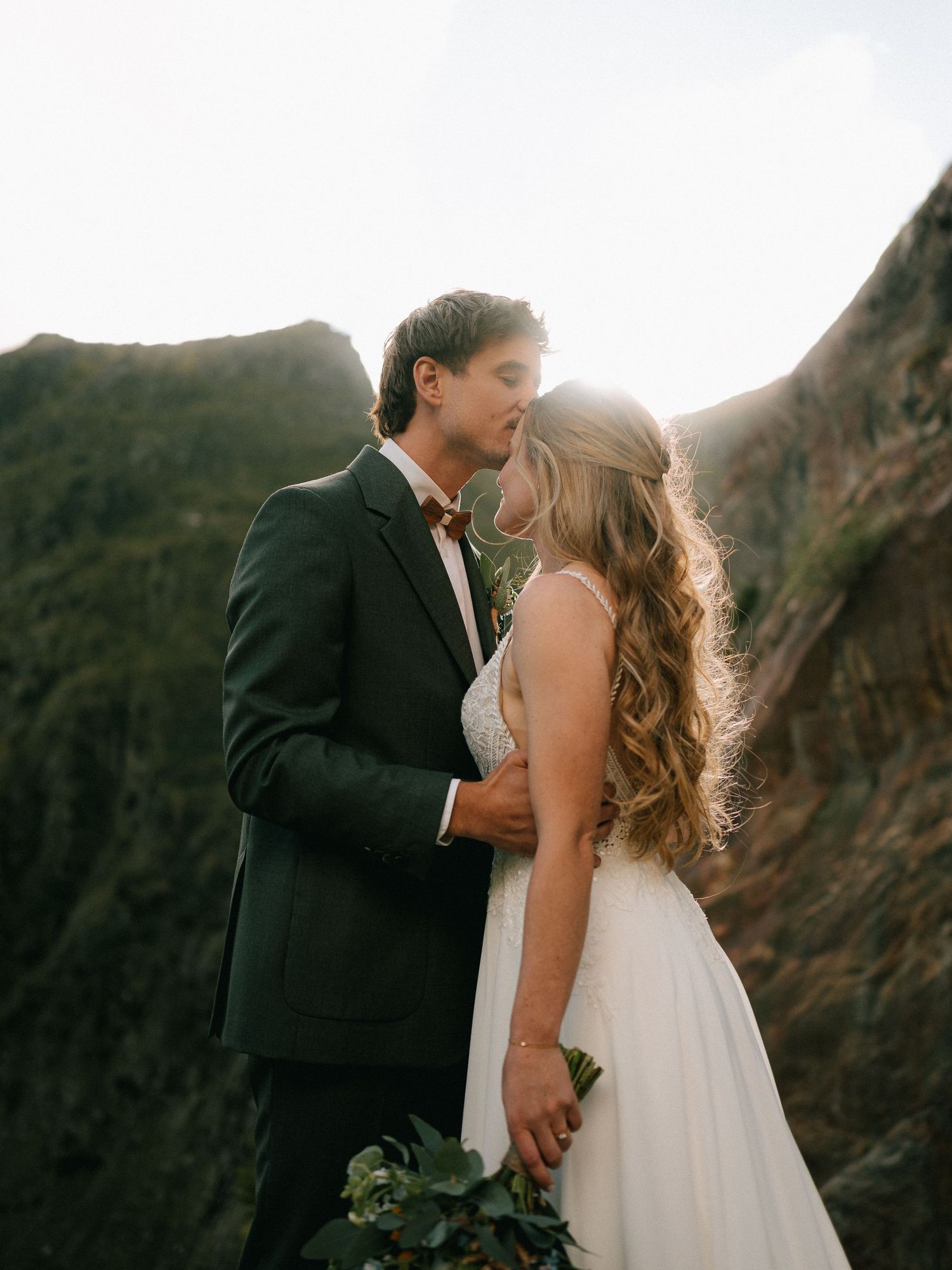 A couple in wedding attire share romantic moments against a dramatic mountain backdrop at sunset.