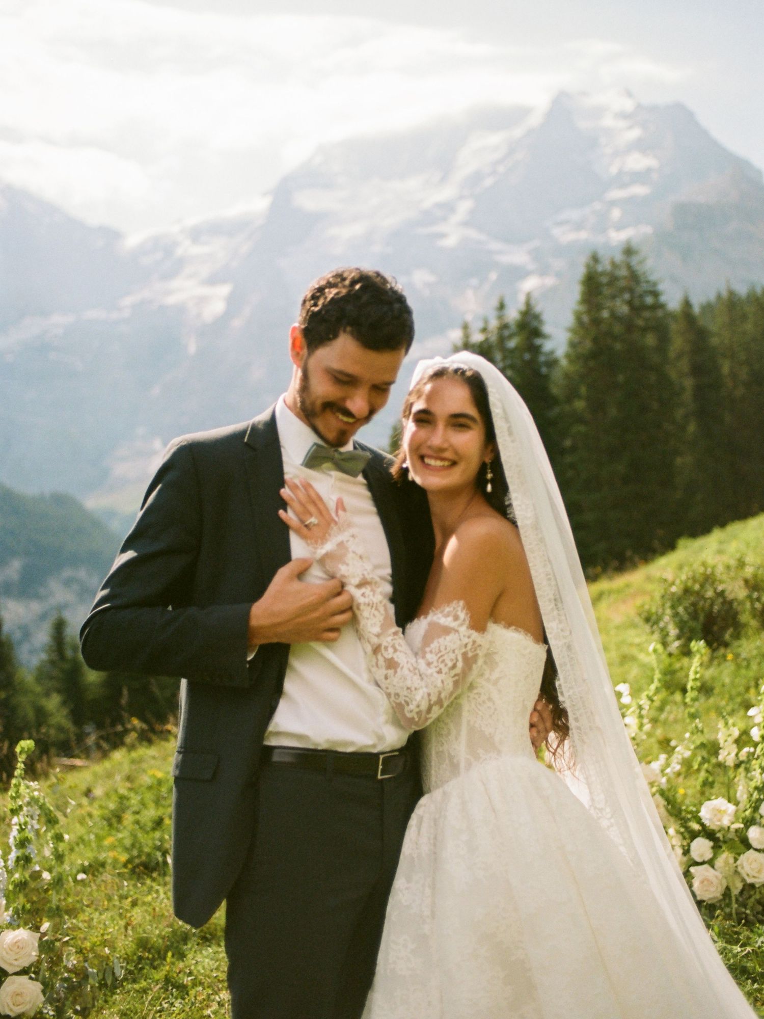 Couple in wedding attire smiling on alpine meadow with snow-capped mountain backdrop on sunny day.