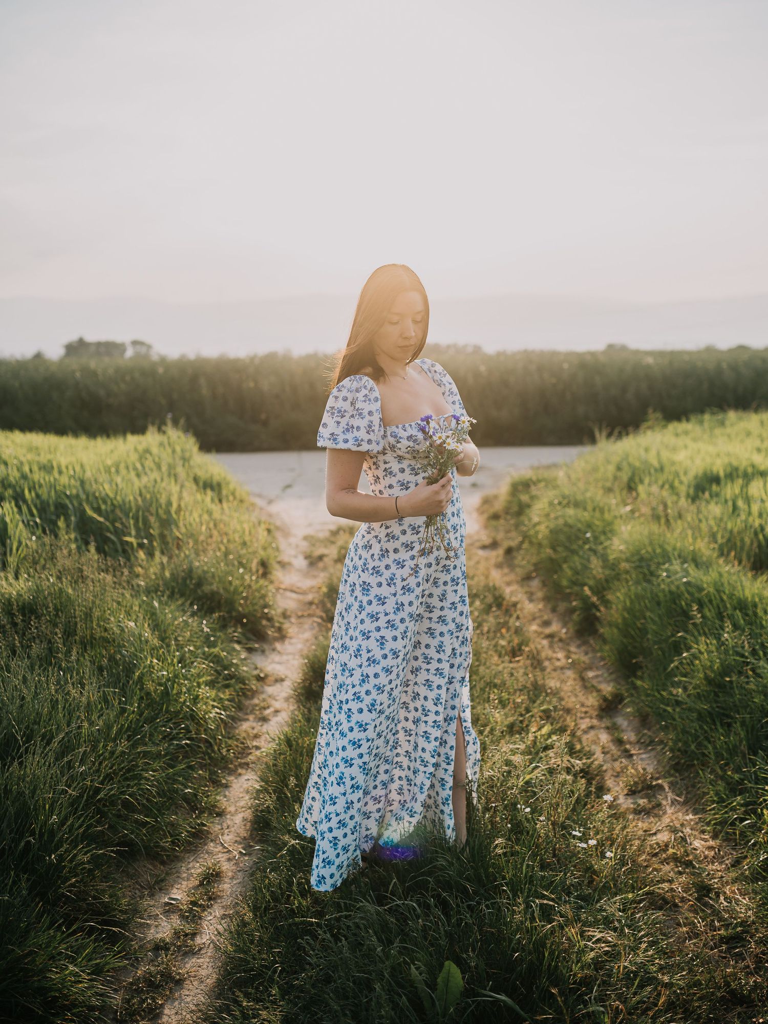 A figure in a flowing blue dress walks along a dirt path through green fields at sunset.