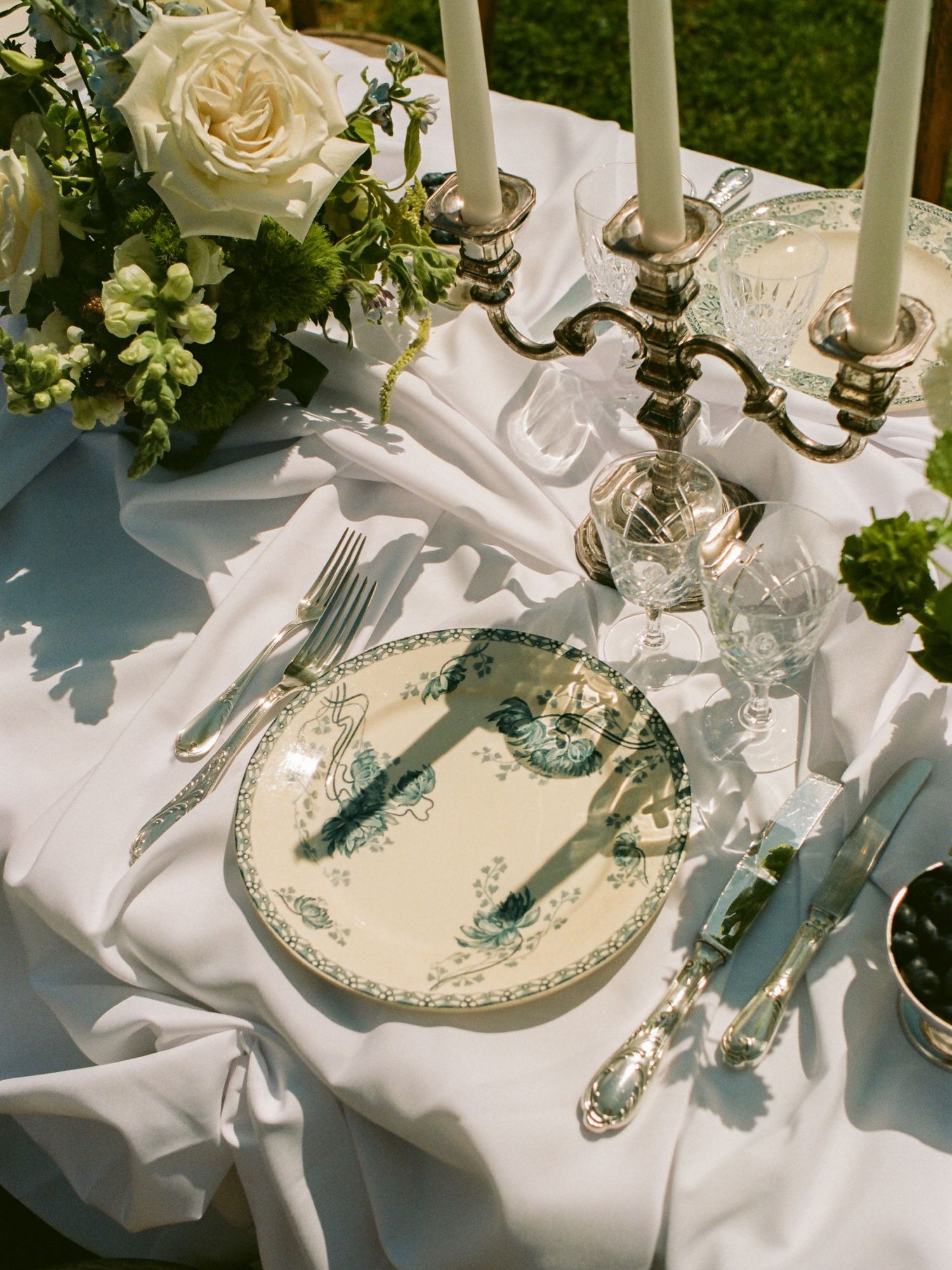 Elegant wedding table setting with floral china, silver candelabra, crystal glasses, and white roses on a draped linen tablecloth.