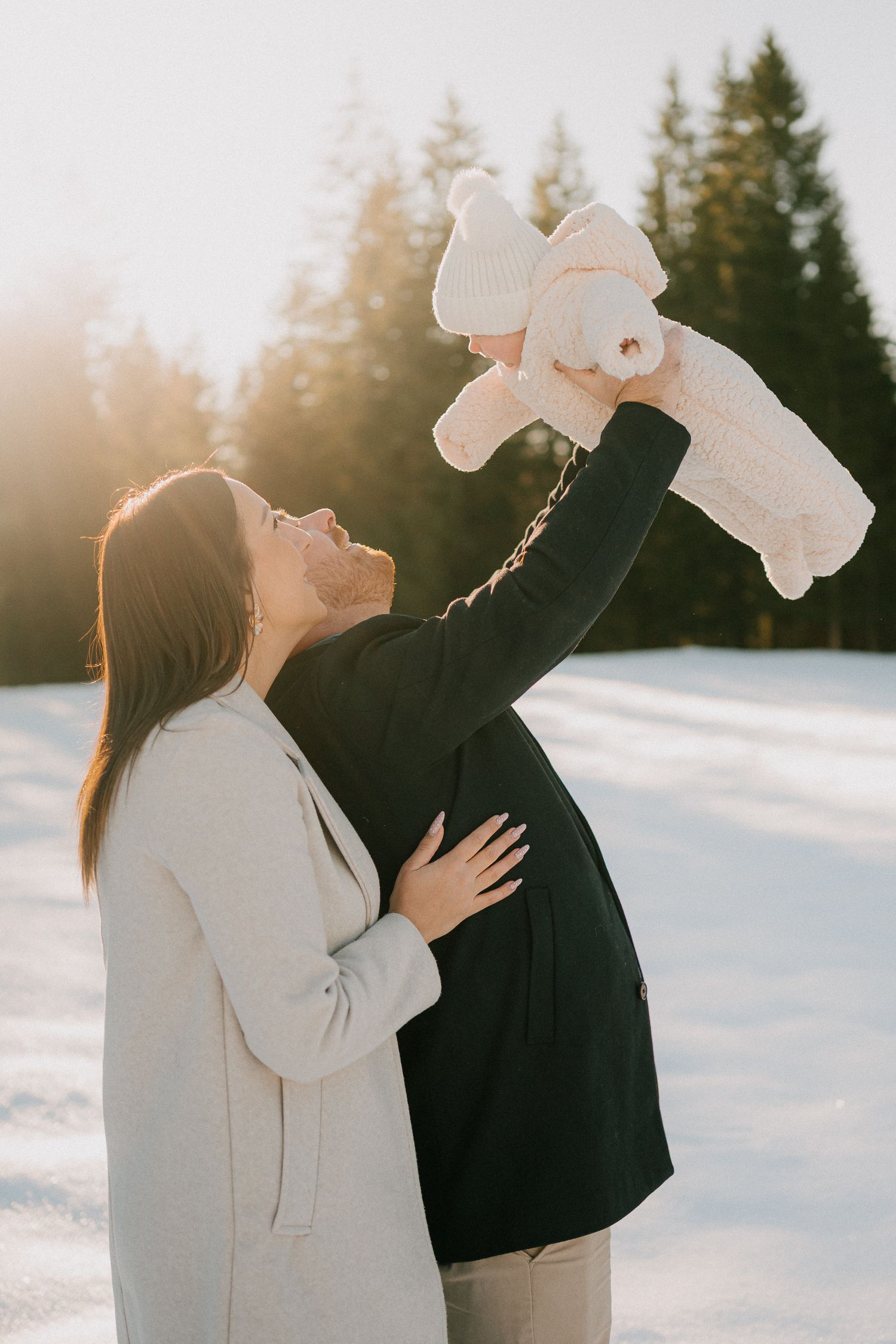 A couple in winter attire plays with a stuffed animal in the snow during golden hour with evergreen trees in background.