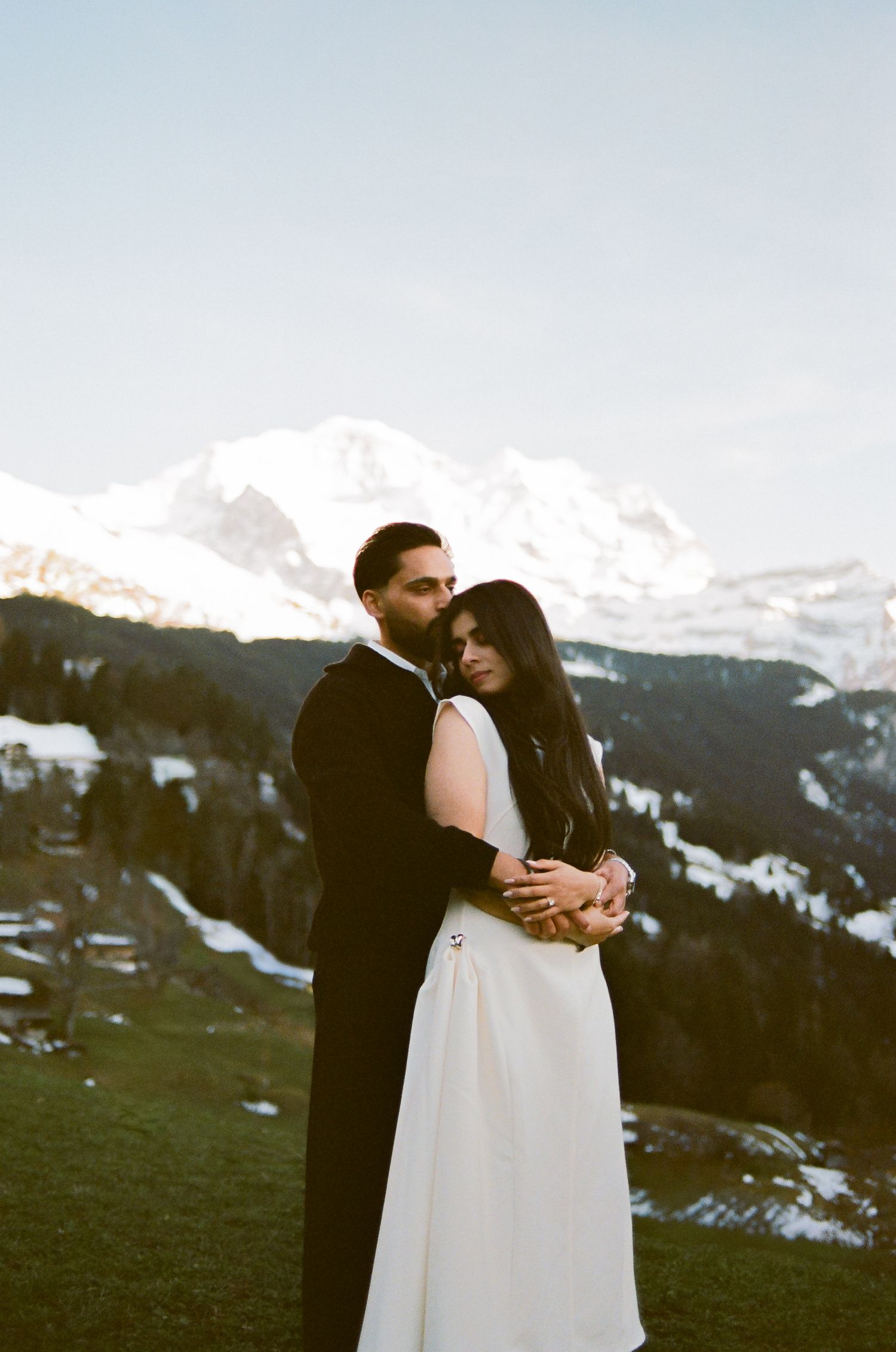 Couple embracing in wedding attire with snow-capped mountain backdrop at golden hour.