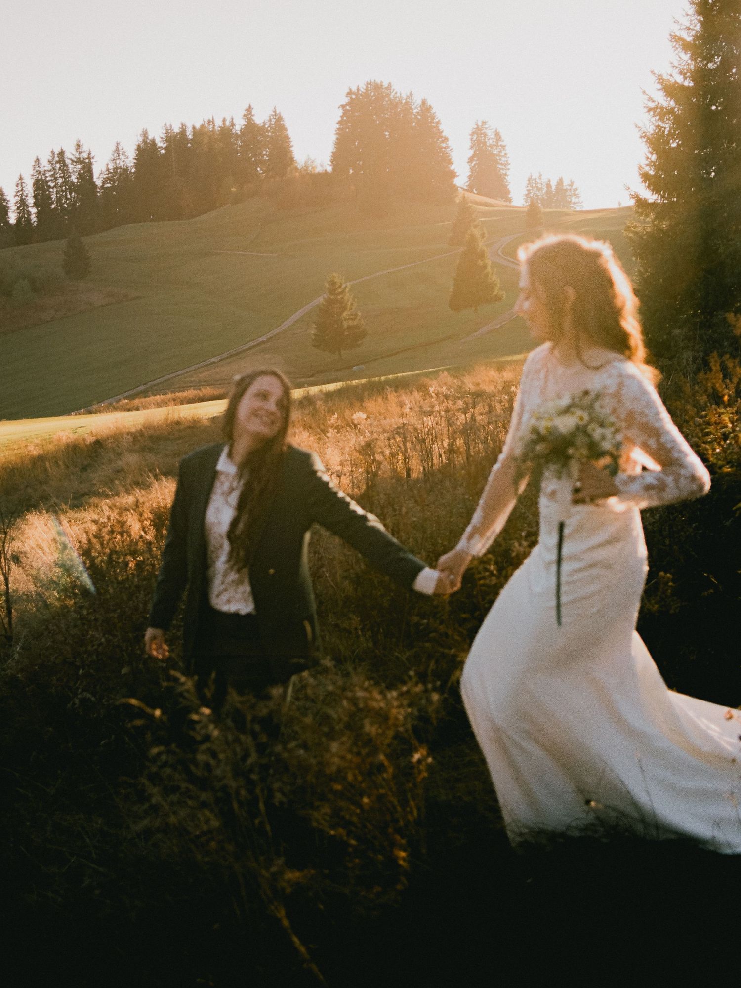 Two brides holding hands in a sunlit meadow, one in a white gown and one in a black suit, at golden hour.