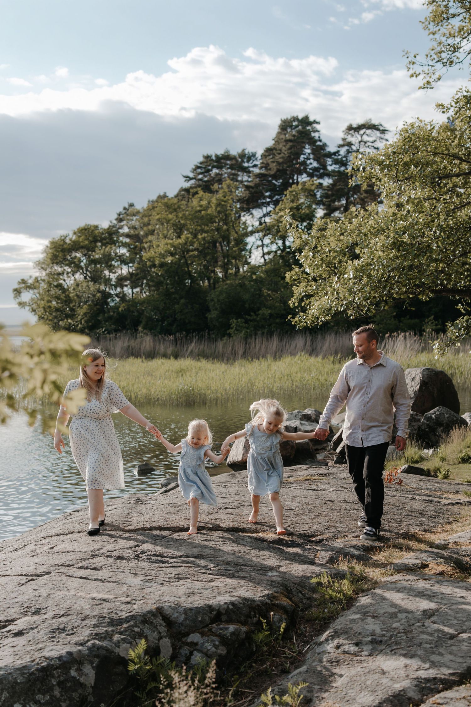 Family of four walking hand-in-hand on rocky lakeside, two girls in matching blue dresses, surrounded by lush greenery.
