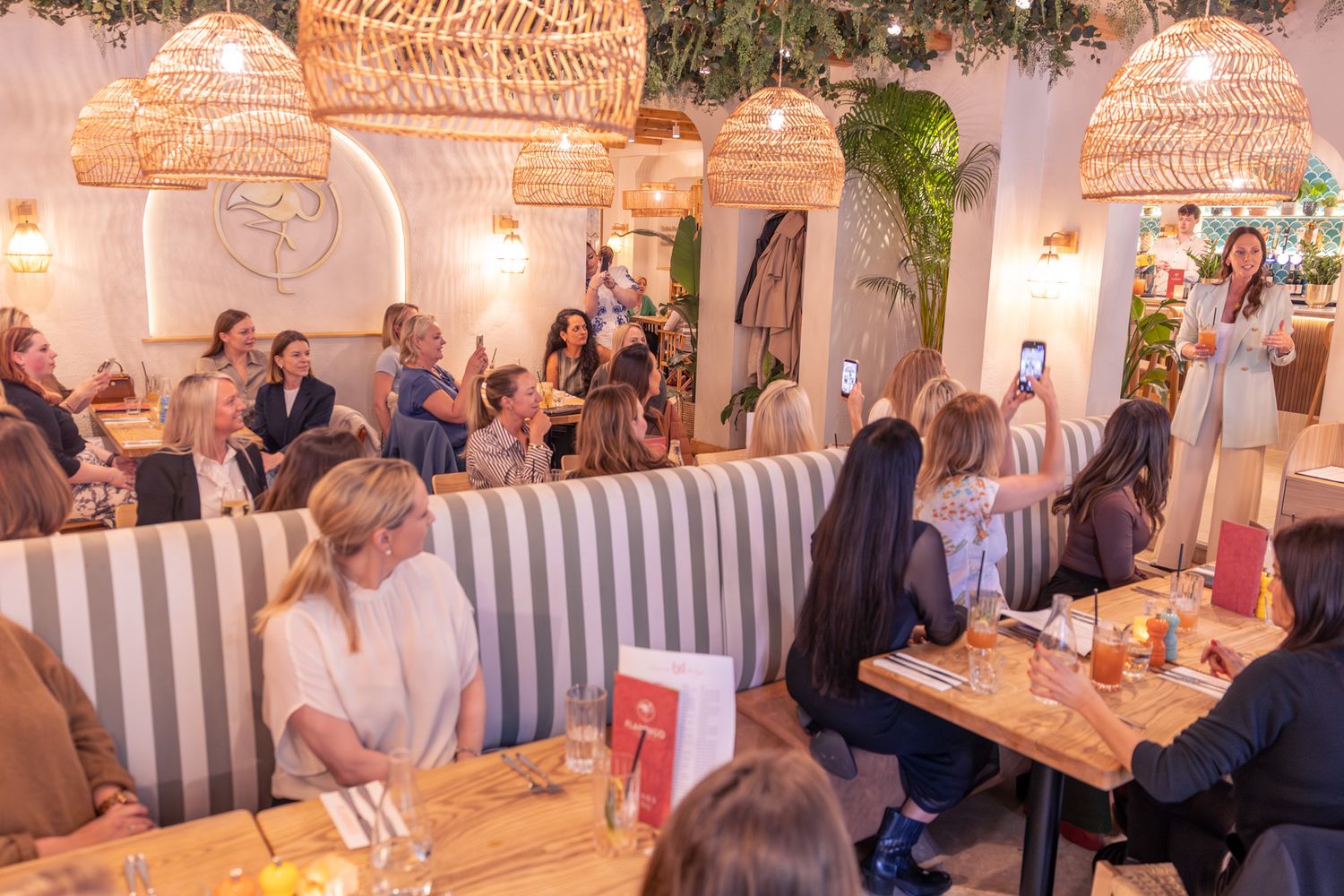 Crowded restaurant with wicker pendant lights, striped booths, and women dining at wooden tables during a social event.