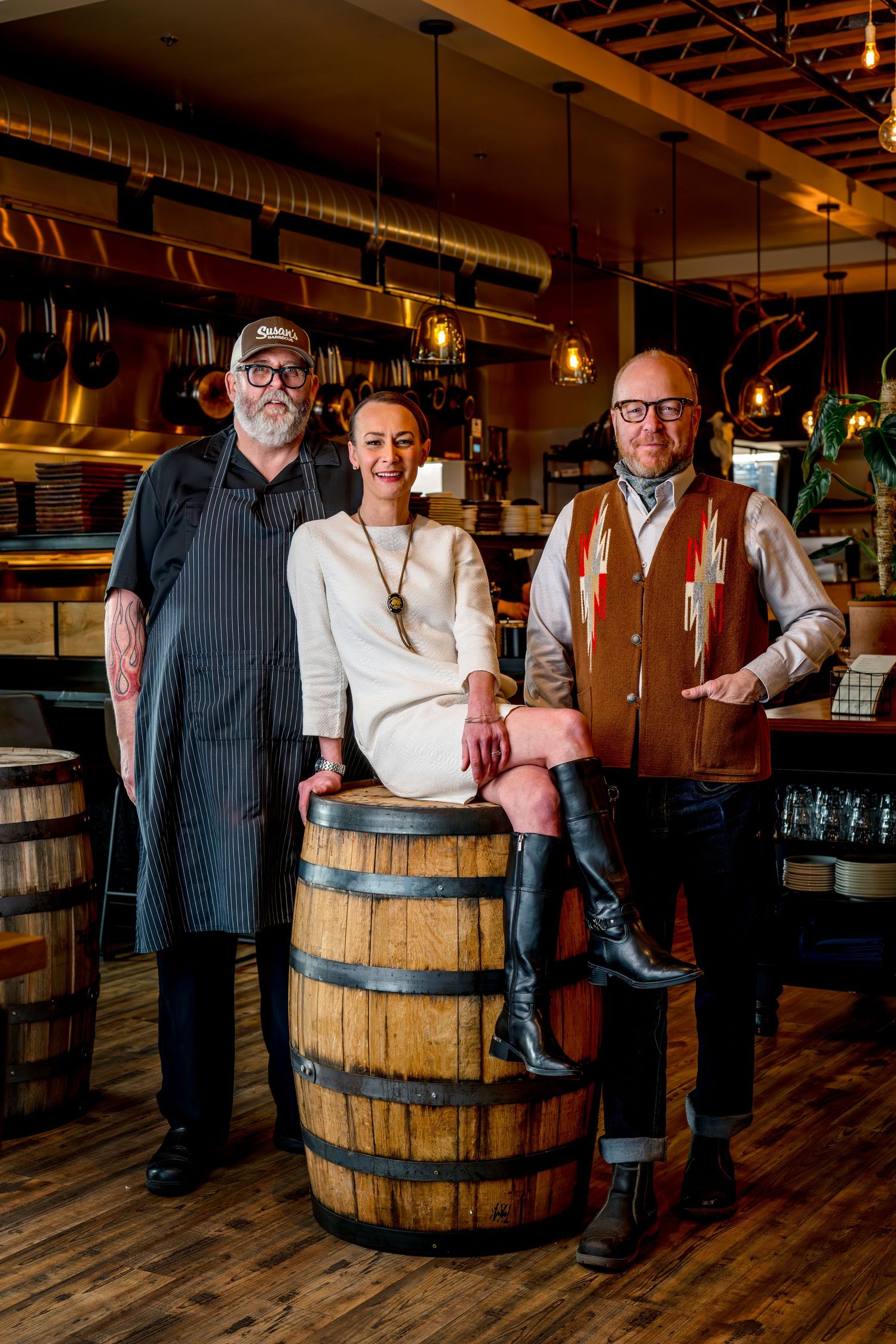 Restaurant staff pose together with wooden barrel in rustic dining space with wood floors and exposed ceiling ducts.