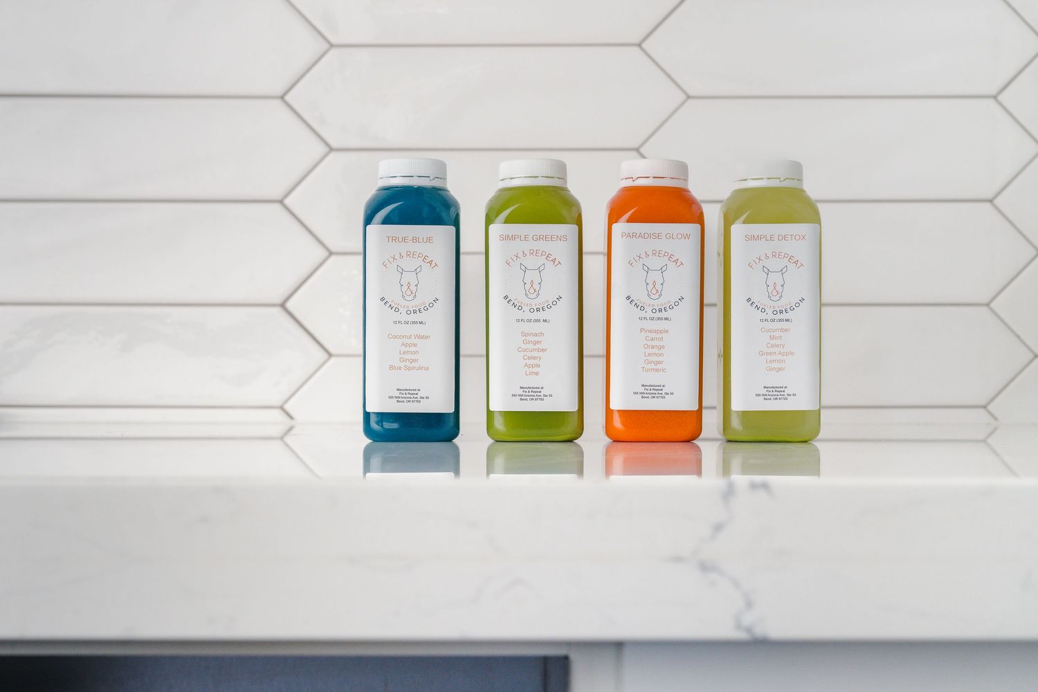 Four colorful glass bottles of cold-pressed juice lined up against a white hexagonal tile wall.
