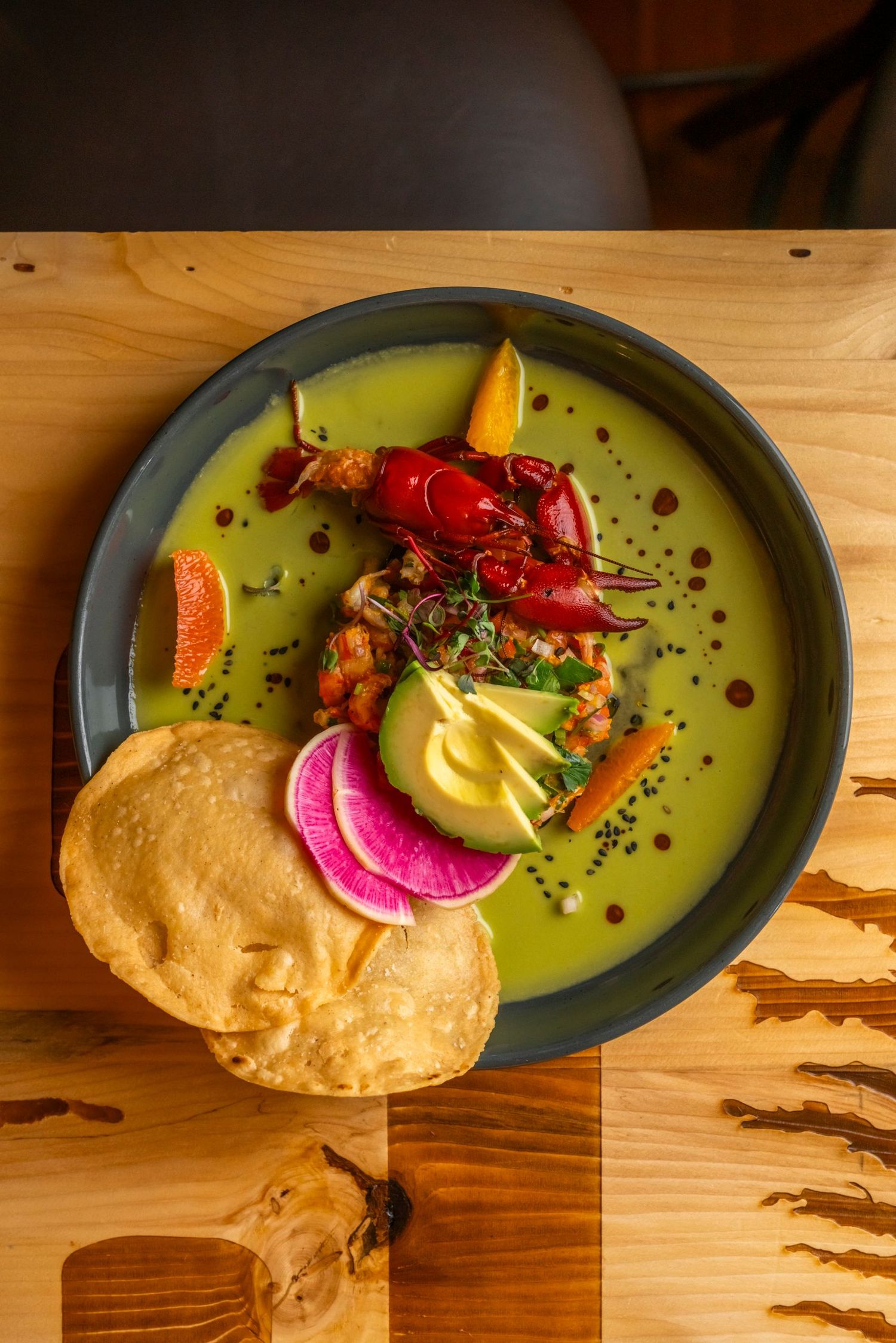 Vibrant green soup served with crispy pappadum, pickled vegetables, and crayfish garnish on rustic wooden table.