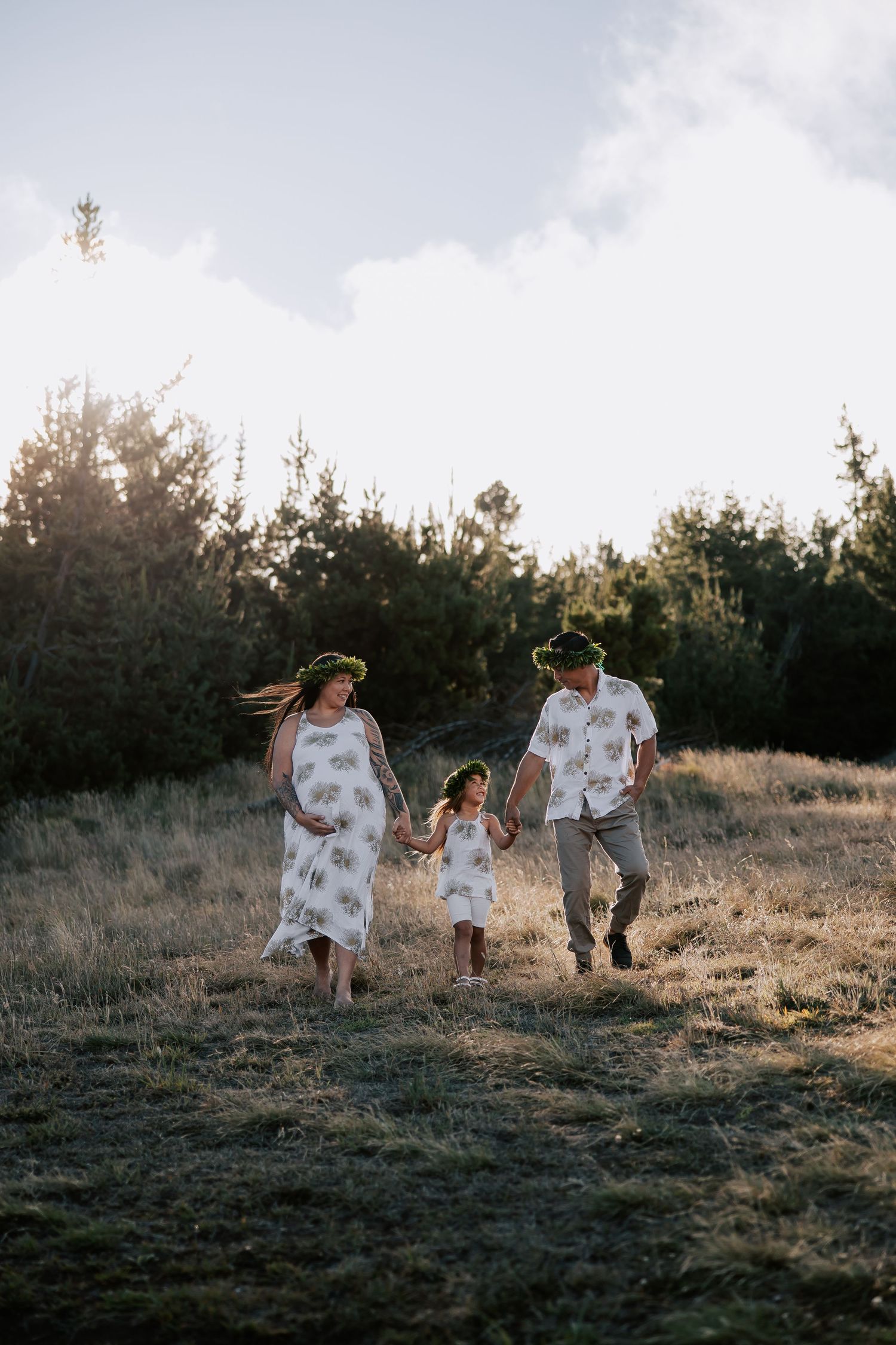Pregnant woman and man in matching floral outfits hold hands with young girl walking through a meadow.