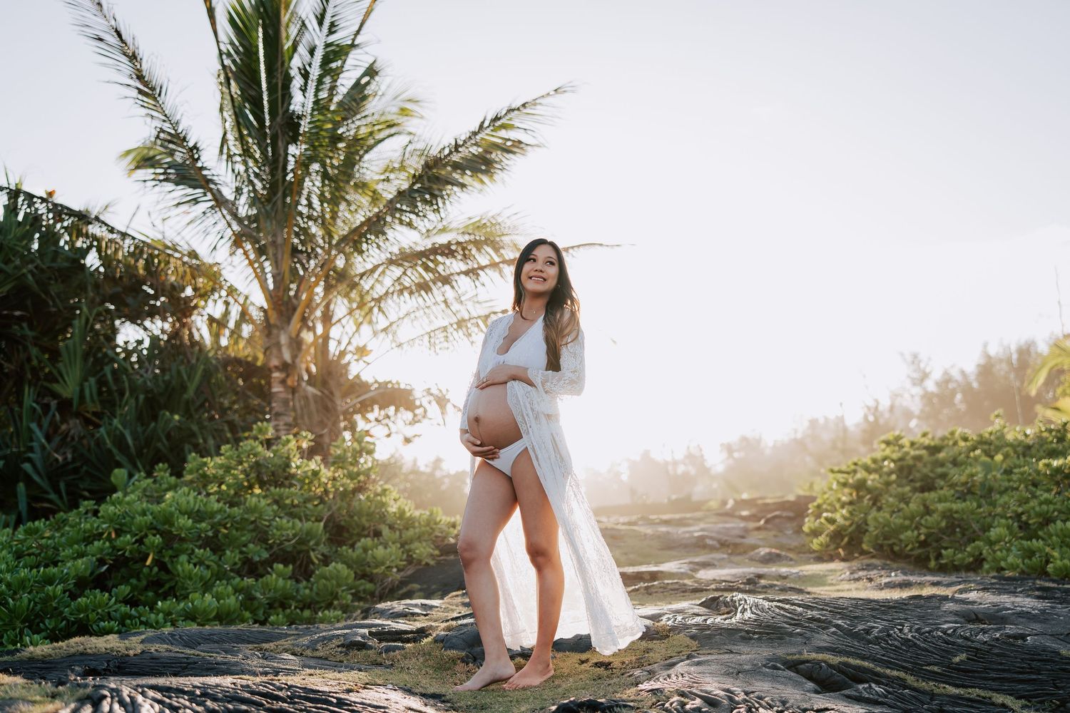 Pregnant woman in white dress standing on rocks with tropical palm trees and golden sunlight in background.