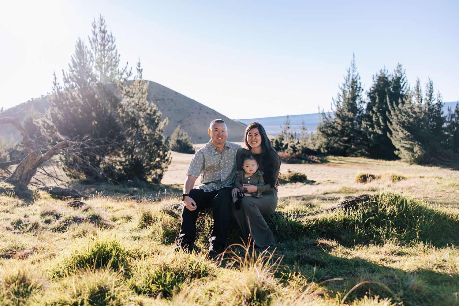 Family of three posing outdoors on a sunny hillside with pine trees and mountains in the background.