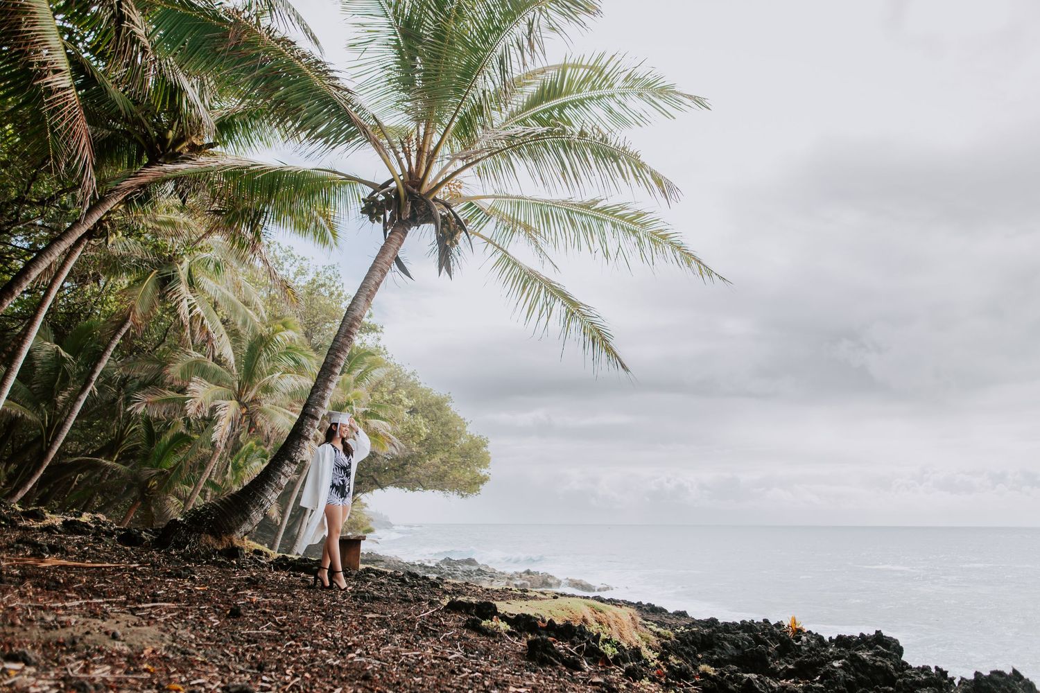 Couple embracing under a leaning palm tree on a rocky Hawaiian coastline with ocean views.