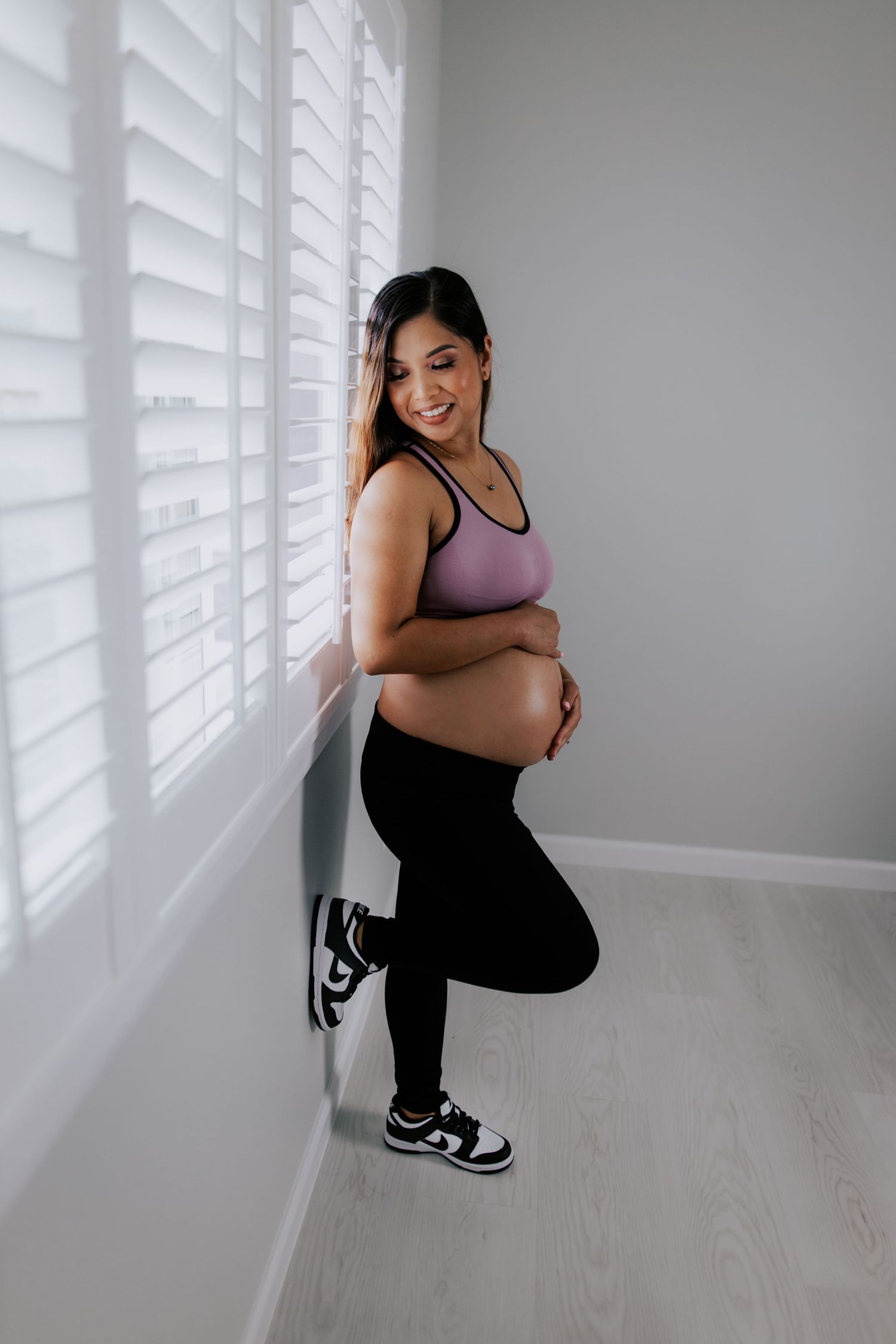 Smiling pregnant woman in purple sports bra and black leggings posing by window shutters in black and white sneakers.