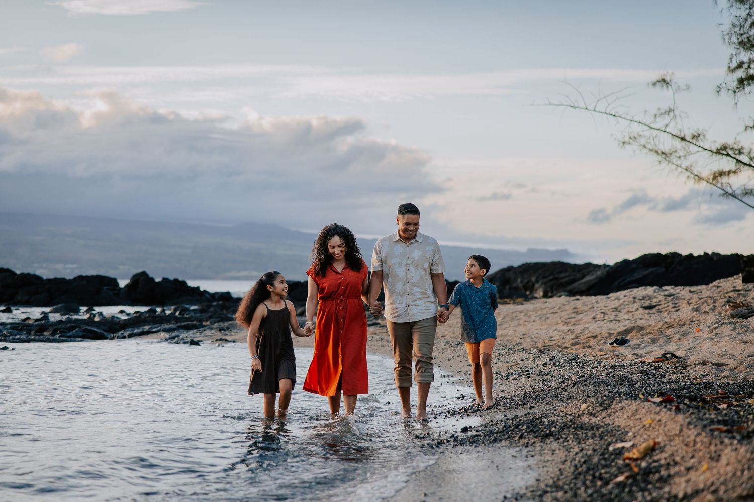 Family of four walks along rocky Hawaiian beach at sunset, parents holding hands with two children in shallow water.