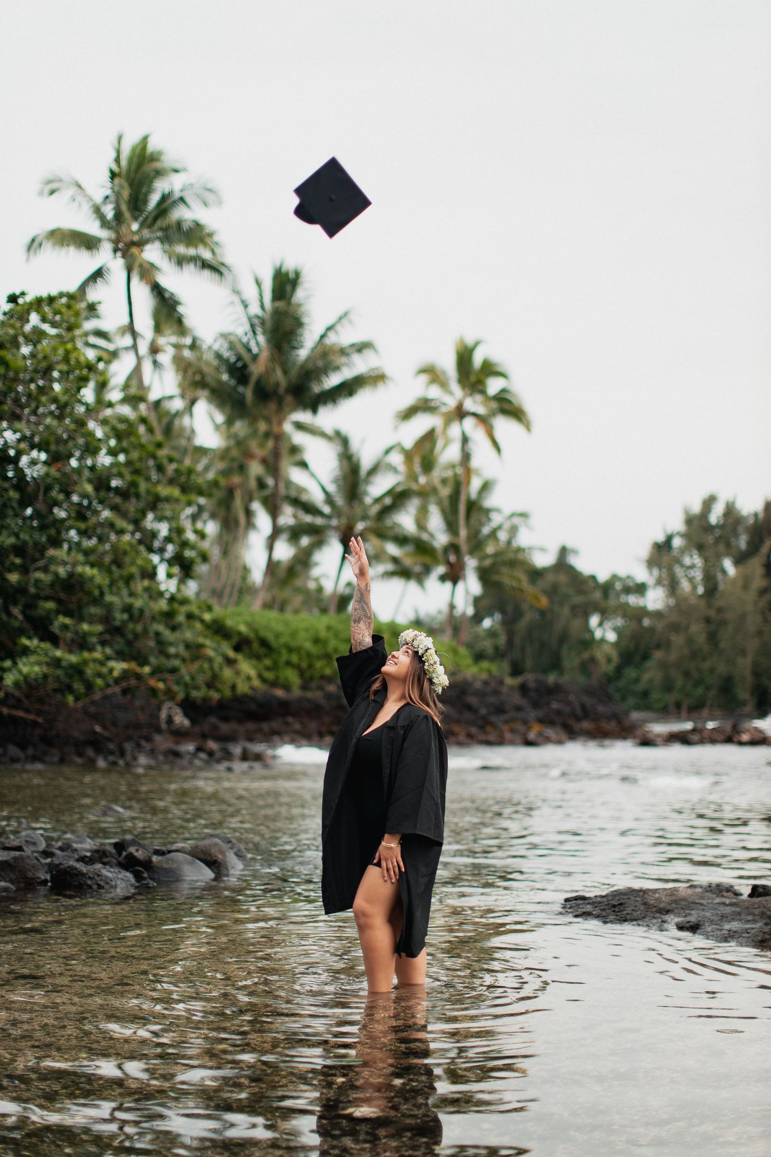 Graduate in black gown tosses cap while standing in tropical ocean waters surrounded by palm trees.