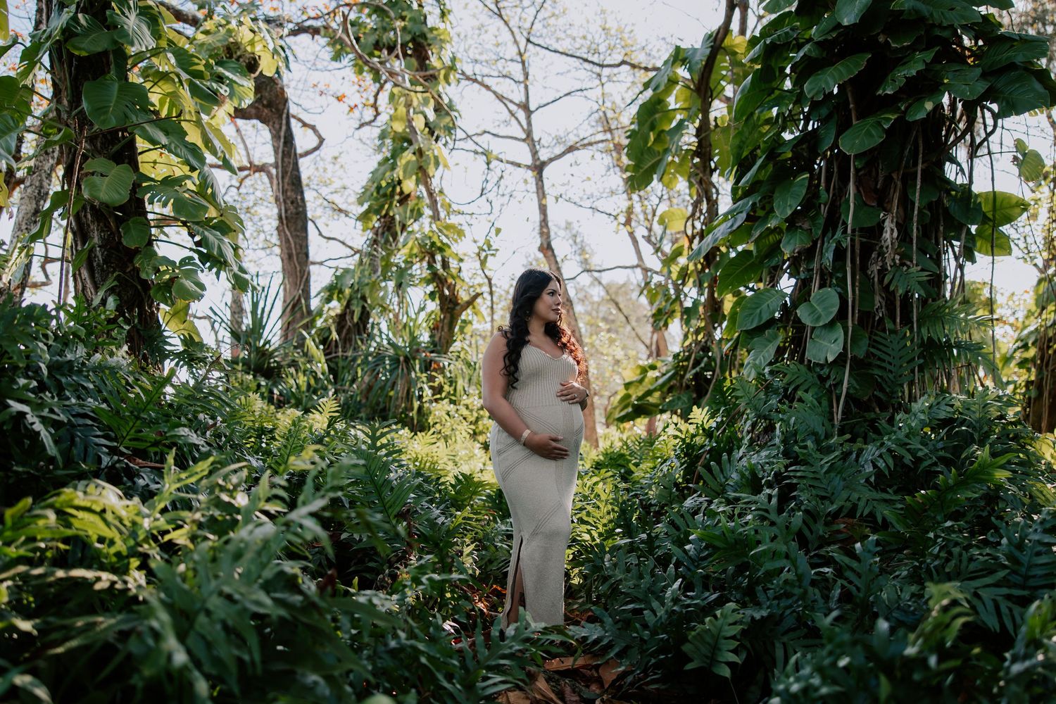 Pregnant woman in white dress standing among lush tropical greenery during an outdoor maternity photoshoot.