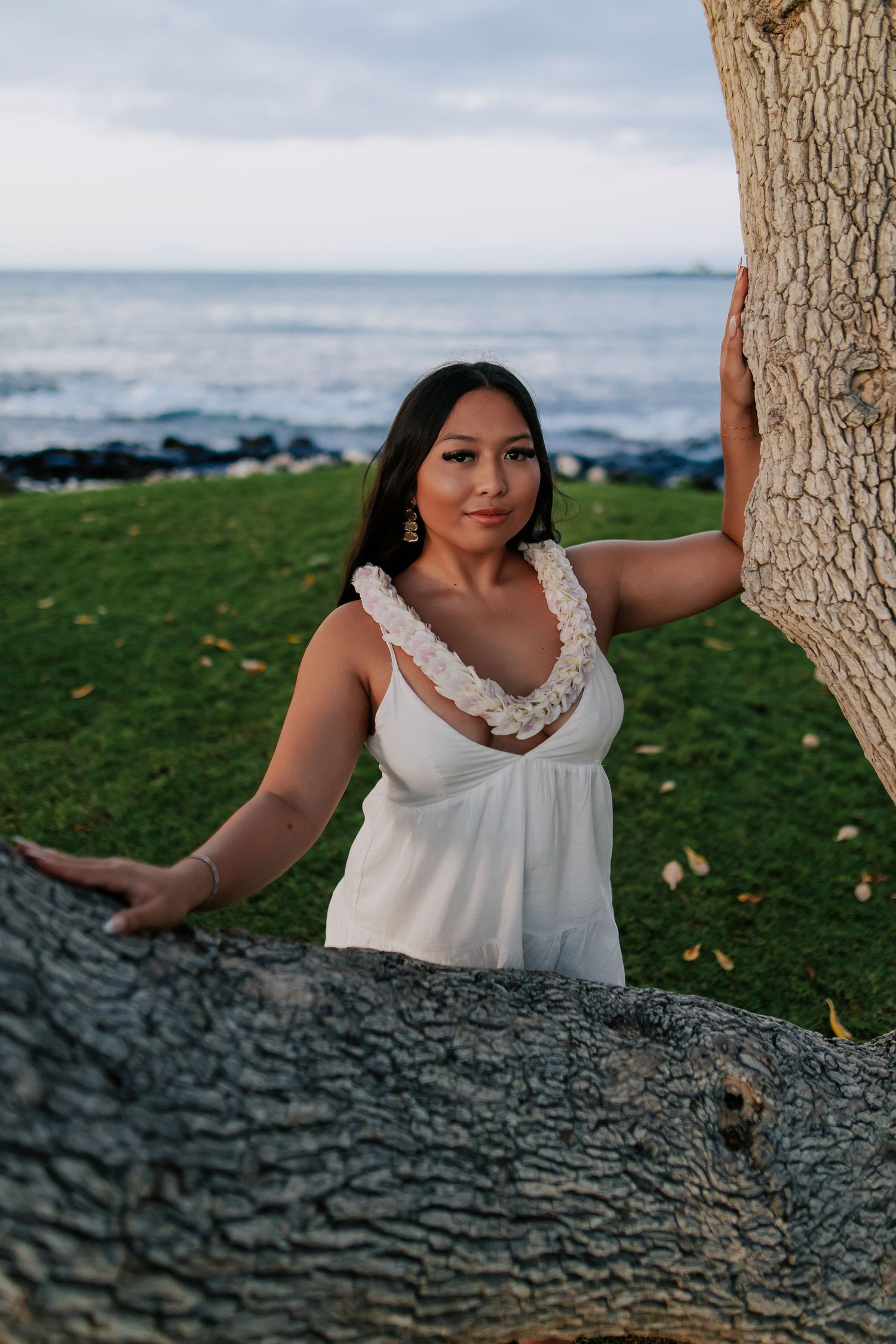 Young woman in white dress poses between palm trees by rocky Hawaiian coastline at dusk.