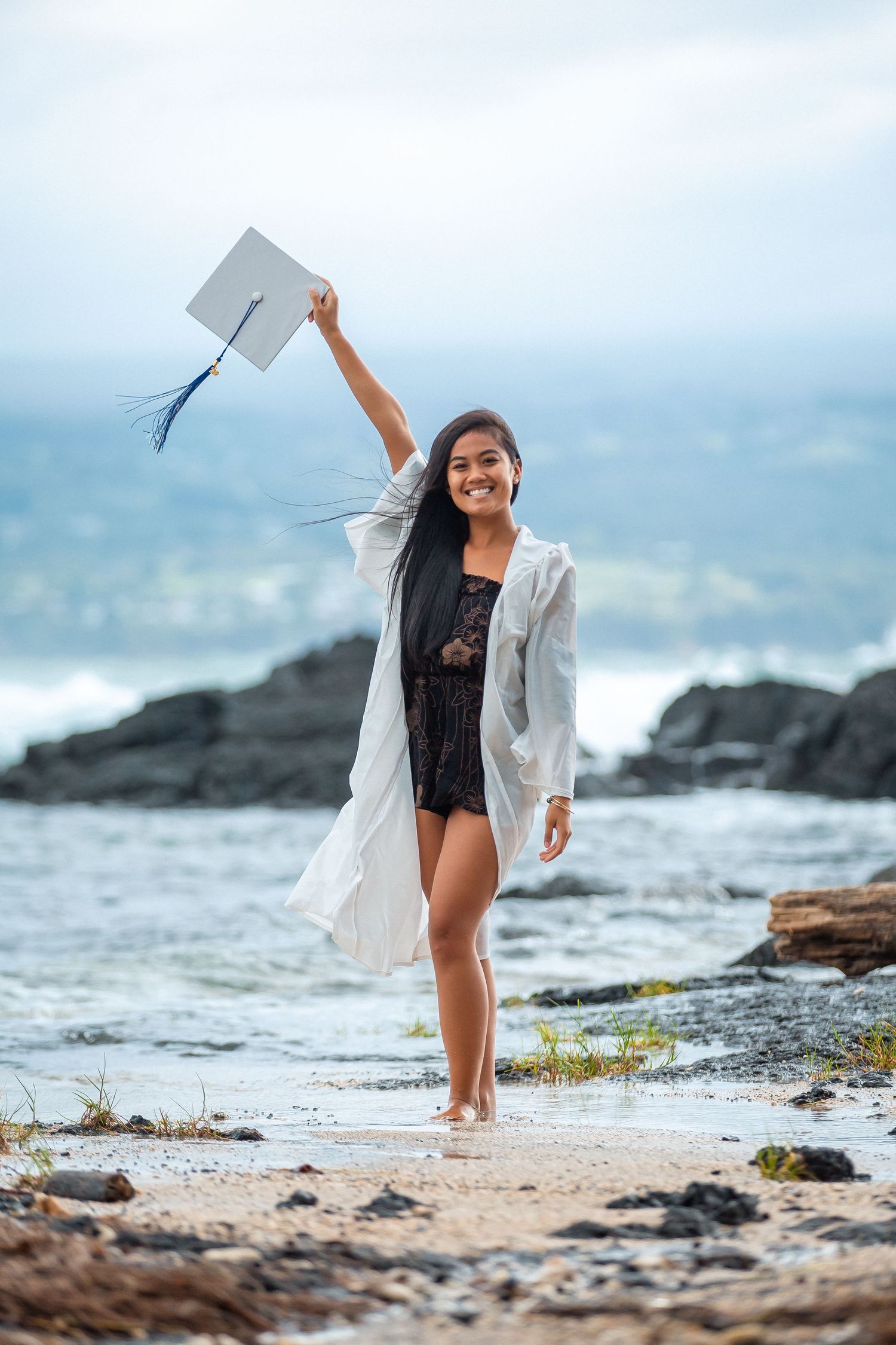 Smiling graduate holds cap aloft on rocky beach wearing white robe, celebrating achievement by the ocean.
