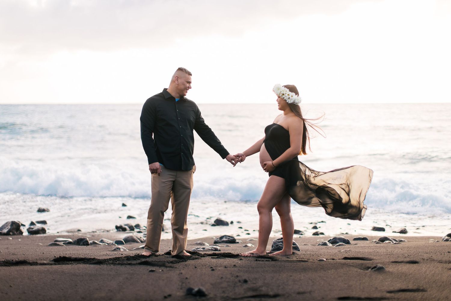 Couple holding hands on a black sand beach at sunset, pregnant woman in black maternity swimsuit with flowing gold fabric.
