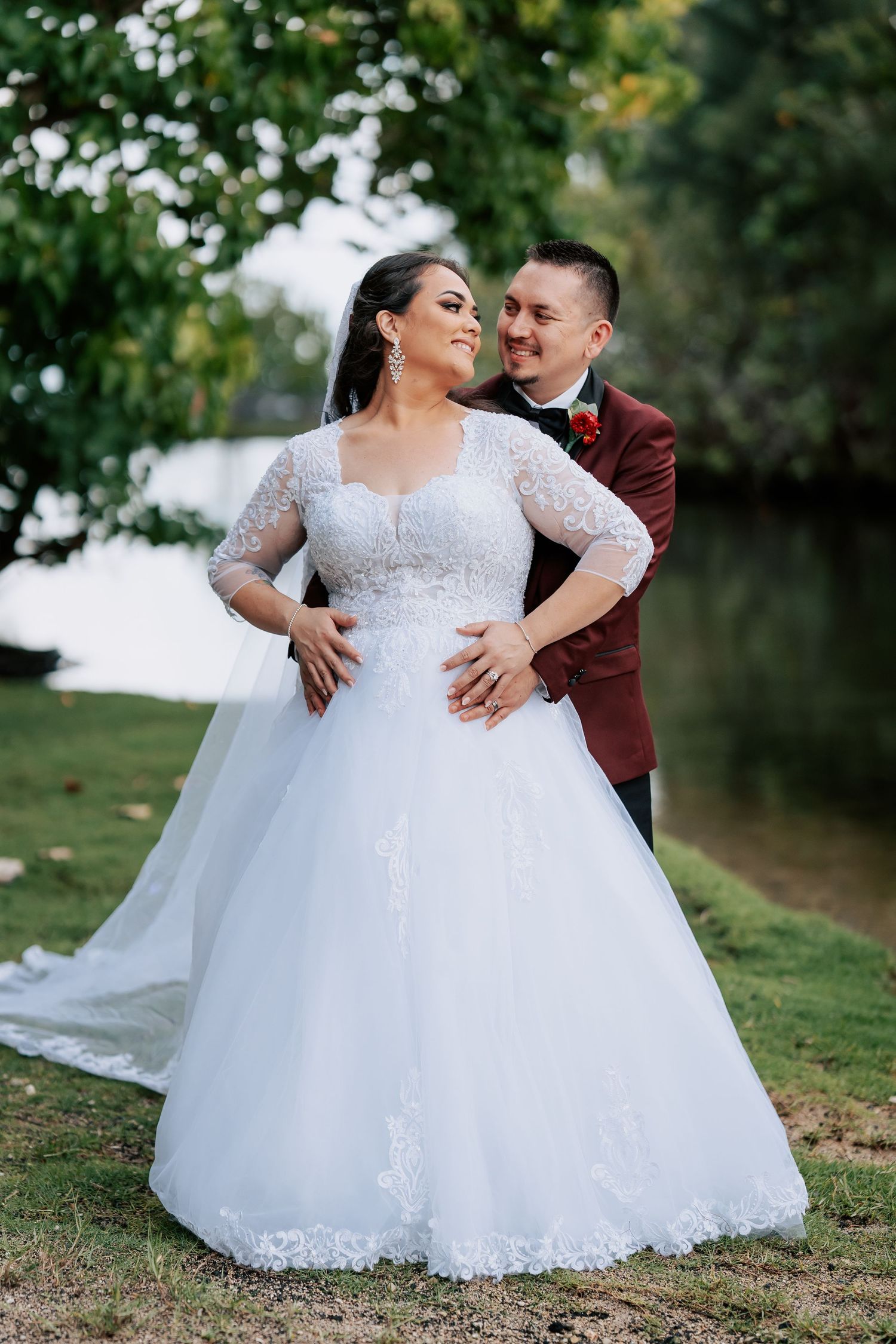 Bride in white lace ball gown with veil stands with groom in burgundy suit outdoors near water.