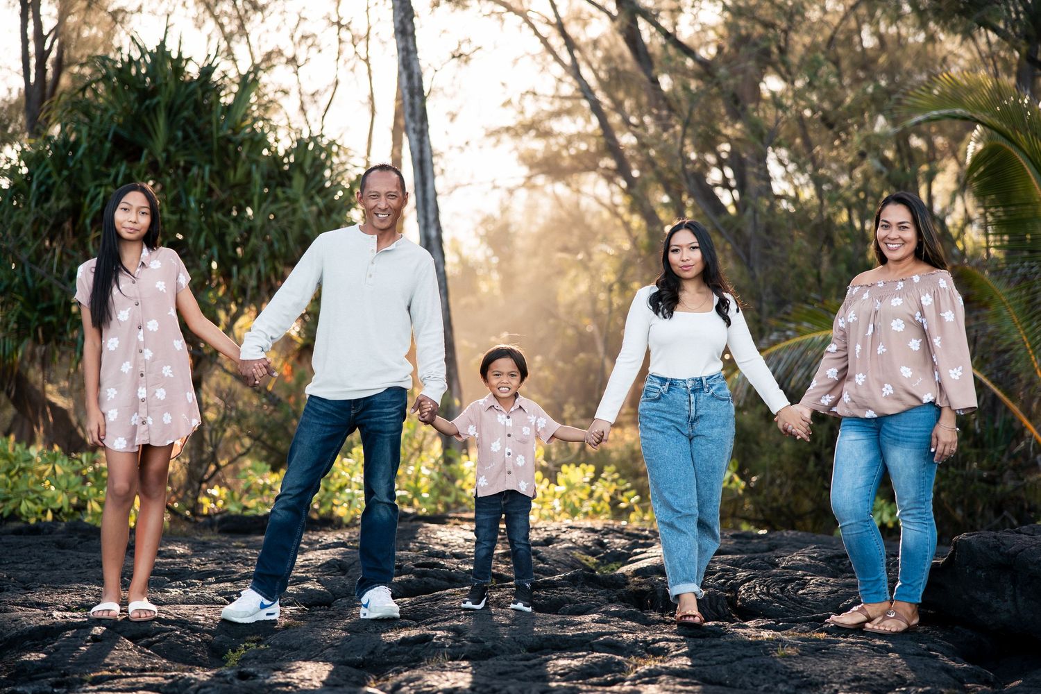 Family of five holding hands outdoors on lava rock, surrounded by lush green trees in warm golden light.