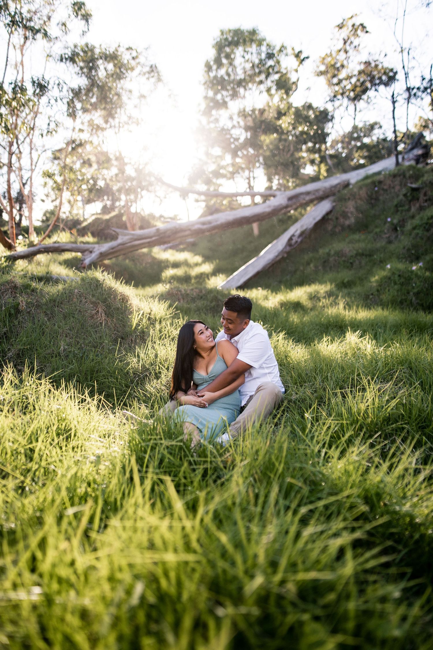 Couple sitting in lush green grass embracing at golden hour with sunlit trees in background.
