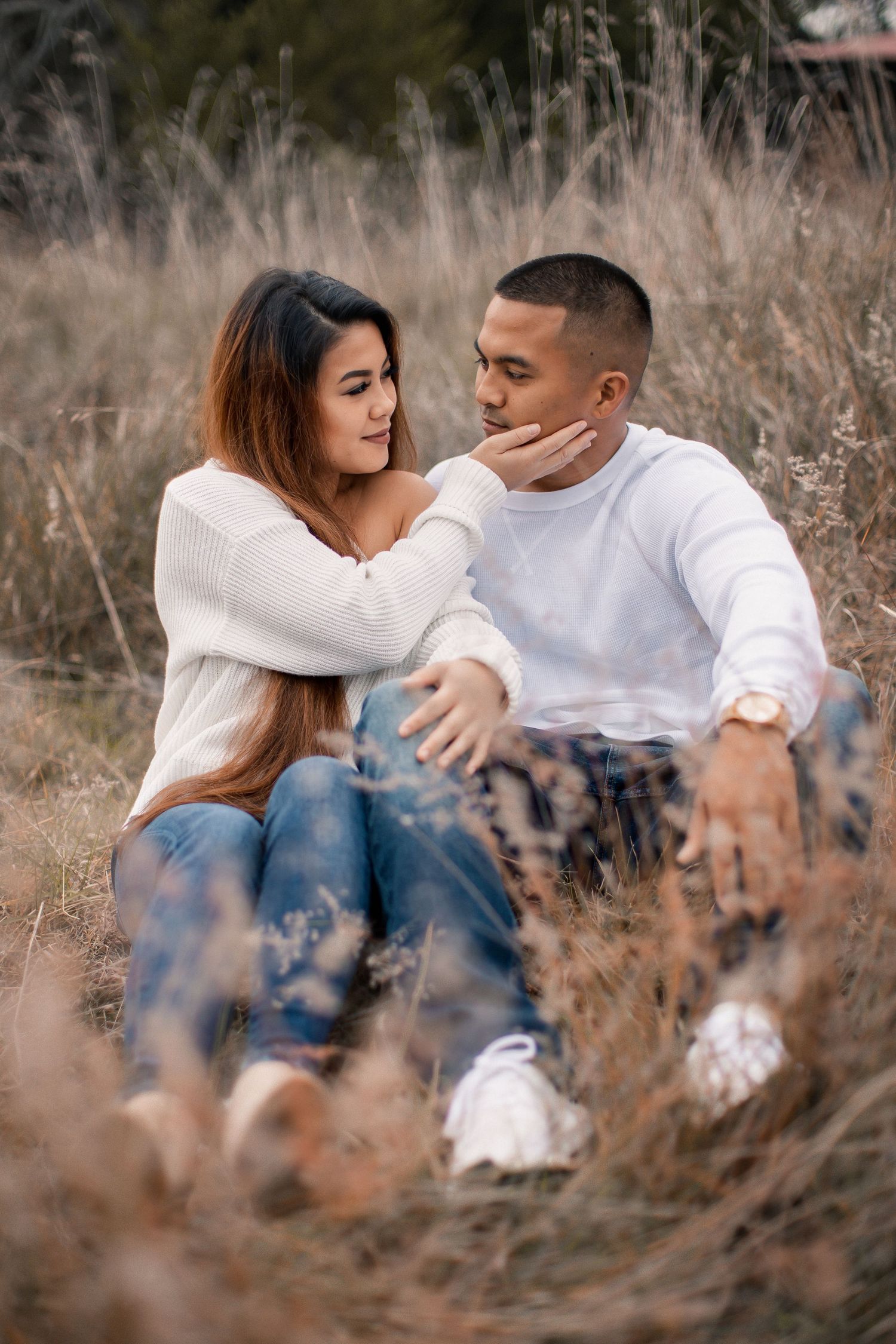 A couple sits romantically in a golden field, woman tenderly touching mans face, both wearing white tops and jeans.