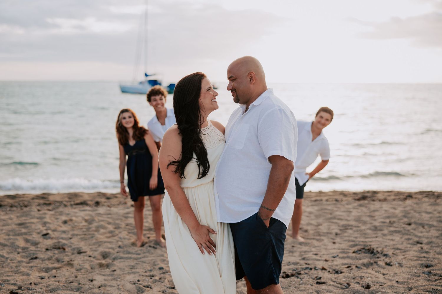 Couple shares romantic moment on beach at sunset with family posing behind them near ocean with sailboat in background.