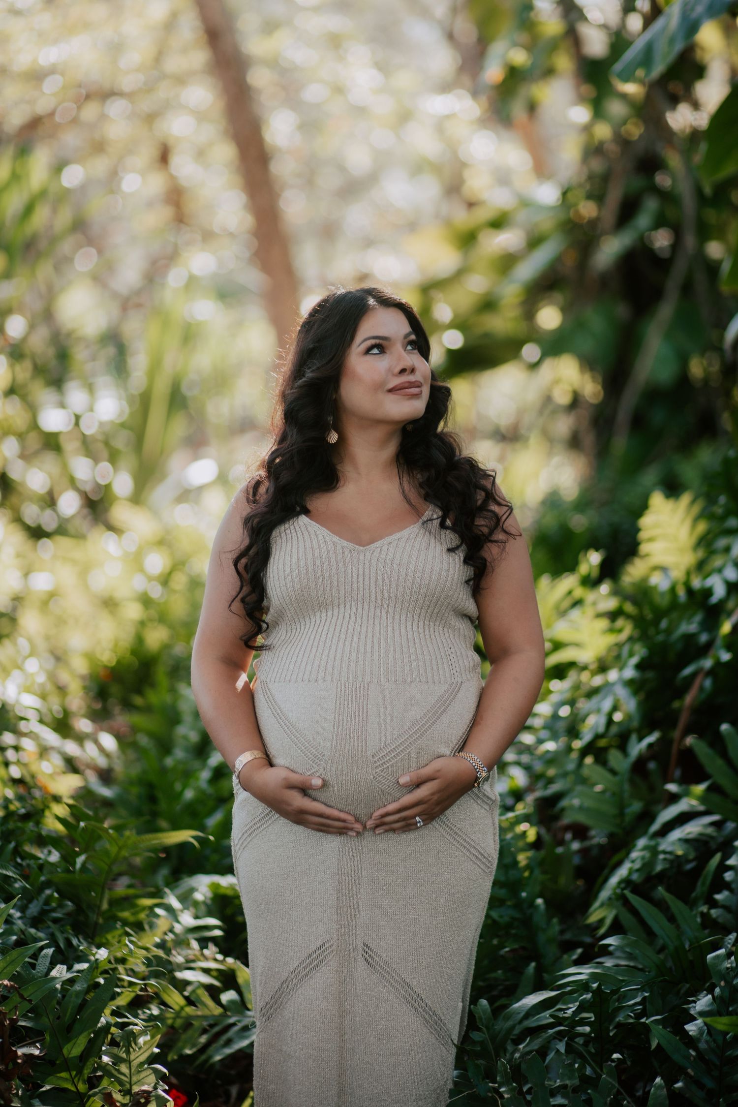 Pregnant woman in beige ribbed dress cradling baby bump, surrounded by lush tropical greenery in a serene outdoor setting.