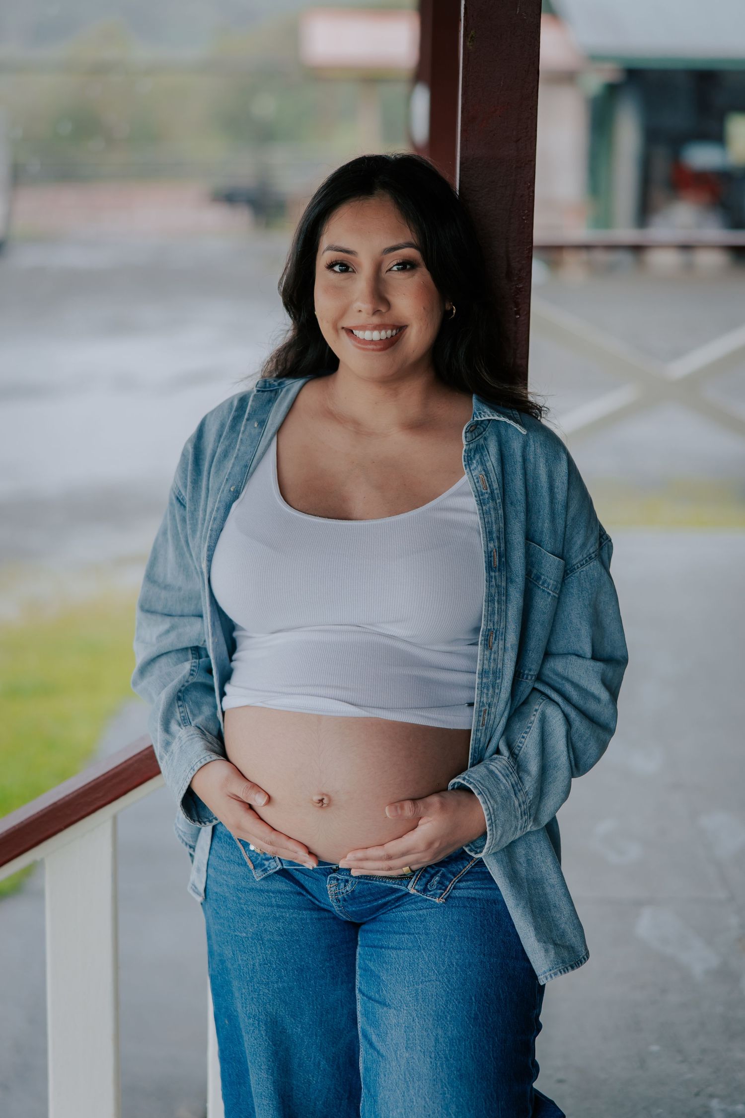 Smiling pregnant woman in white crop top and denim jacket holding her baby bump outdoors on a porch.