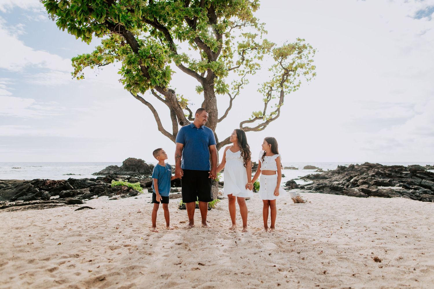 Family of four holding hands on a sandy beach in Hawaii, standing under a tropical tree with rocky shoreline.