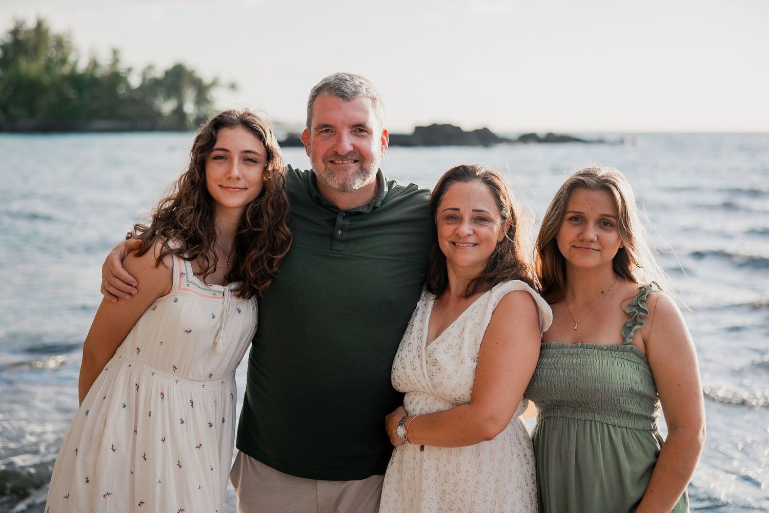 Family of four smiling together on a rocky beach at sunset, wearing casual summer attire.