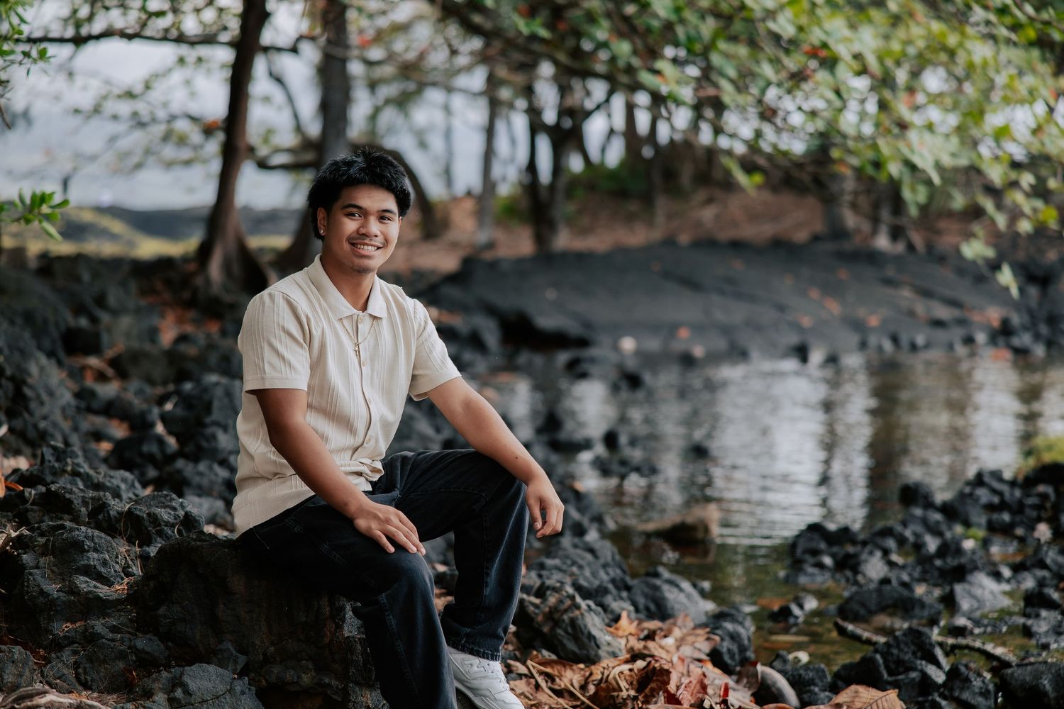 Young man sitting on rocky shoreline near water with trees in background, wearing beige shirt and dark pants.