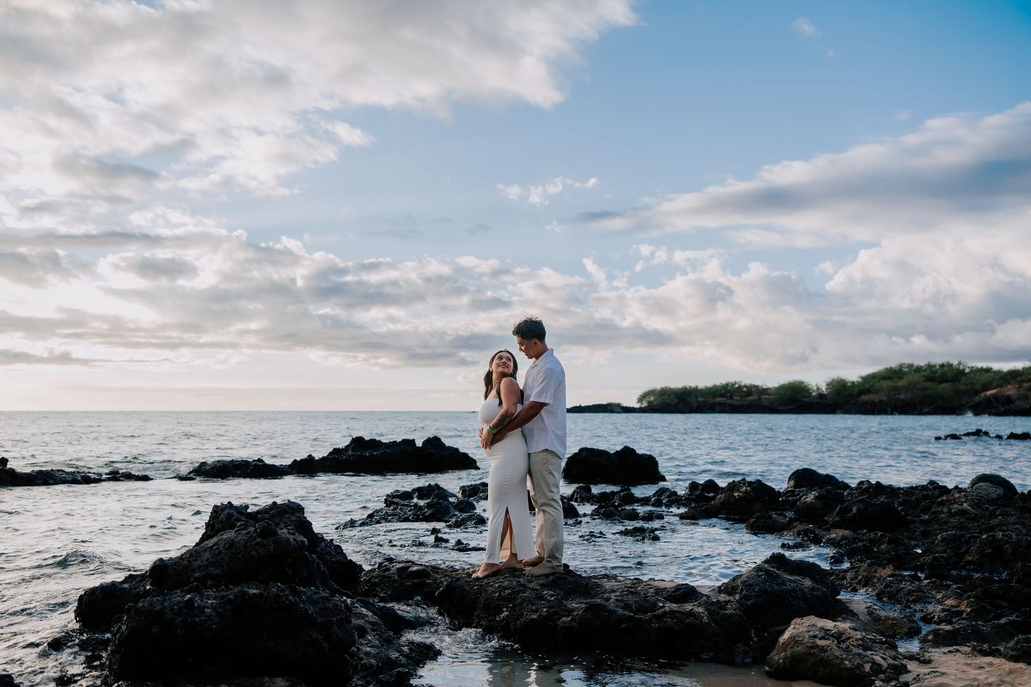 Couple embracing on rocky shoreline at sunset with ocean waves and cloudy sky in Hawaii.