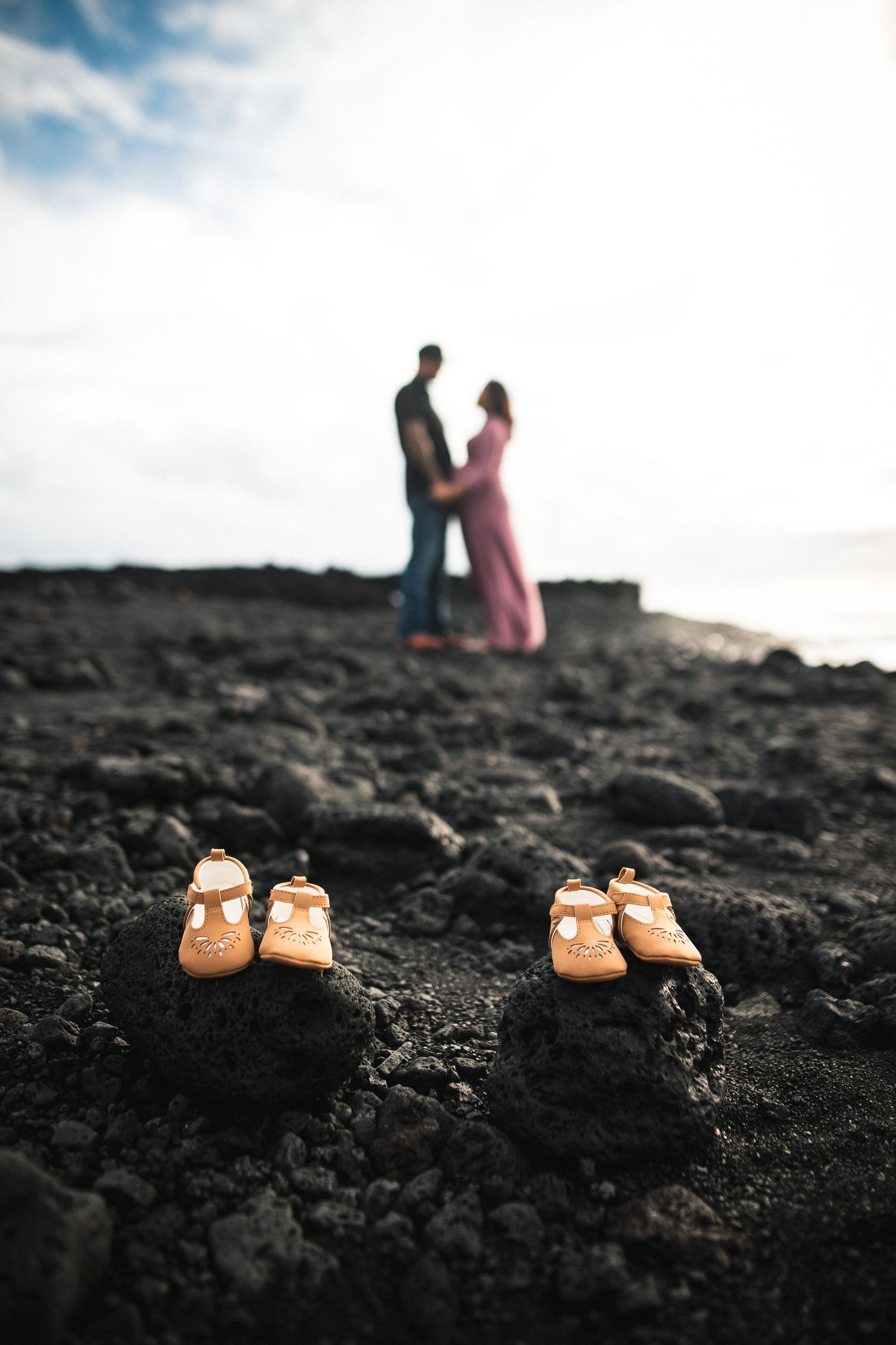 Two pairs of baby shoes on black volcanic rock with a pregnant couple embracing in the blurred background.