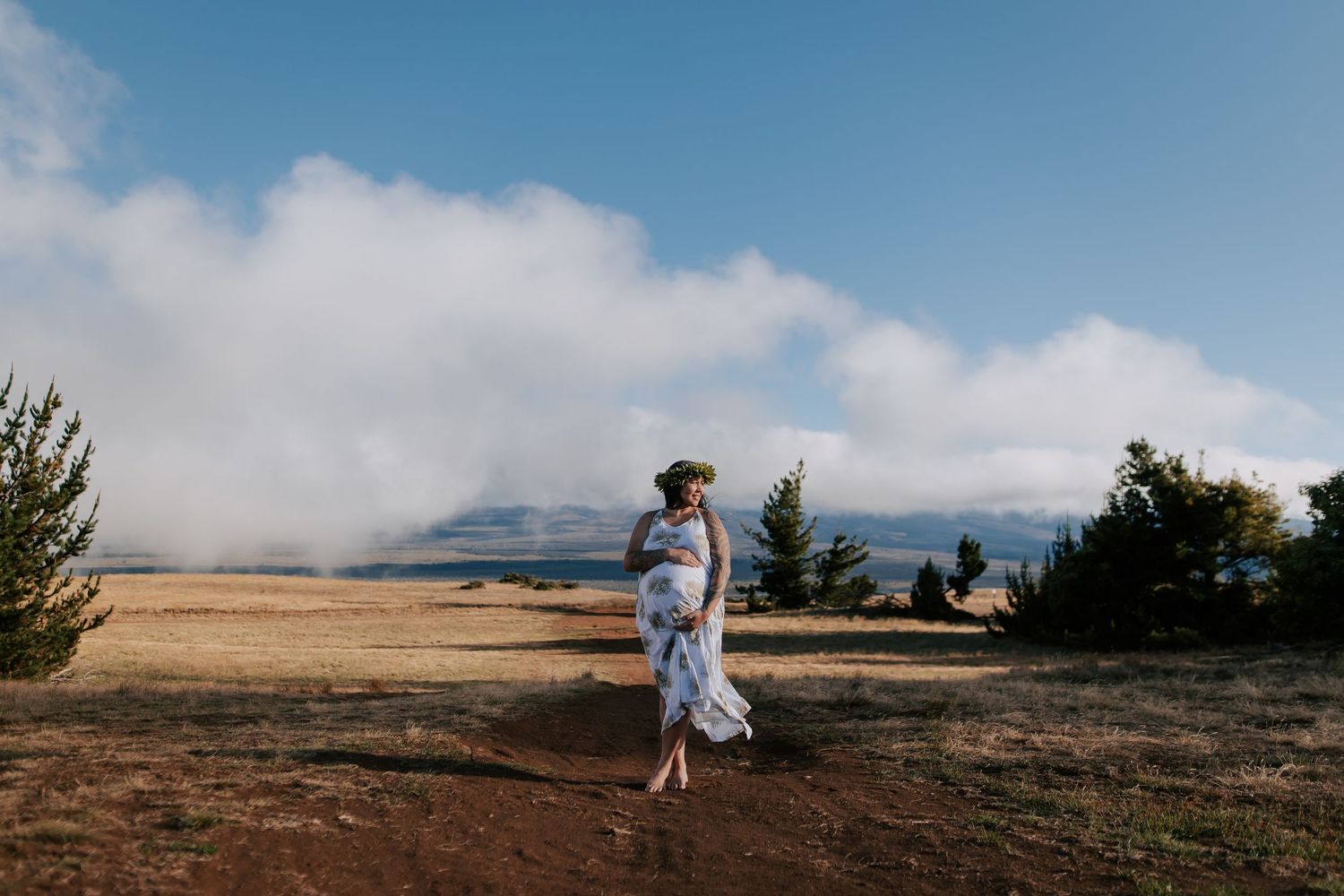Pregnant woman in floral dress walks through scenic mountain landscape under blue sky with clouds.