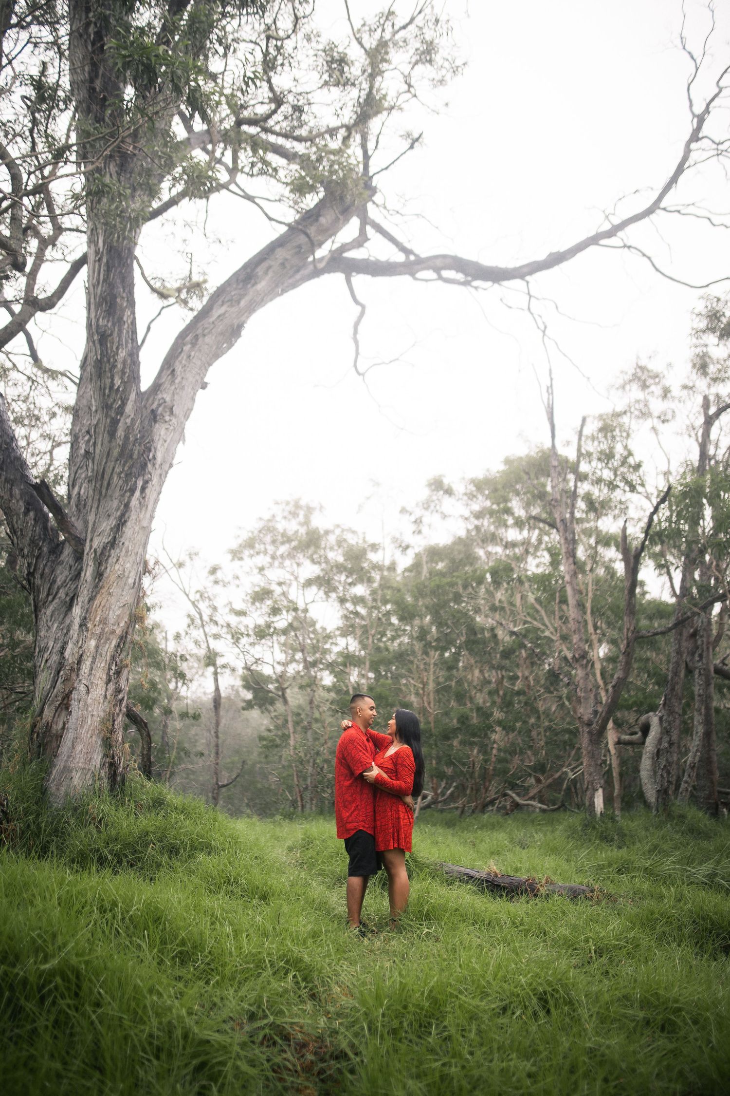 Couple embracing in red outfits among lush green forest trees on a misty day.
