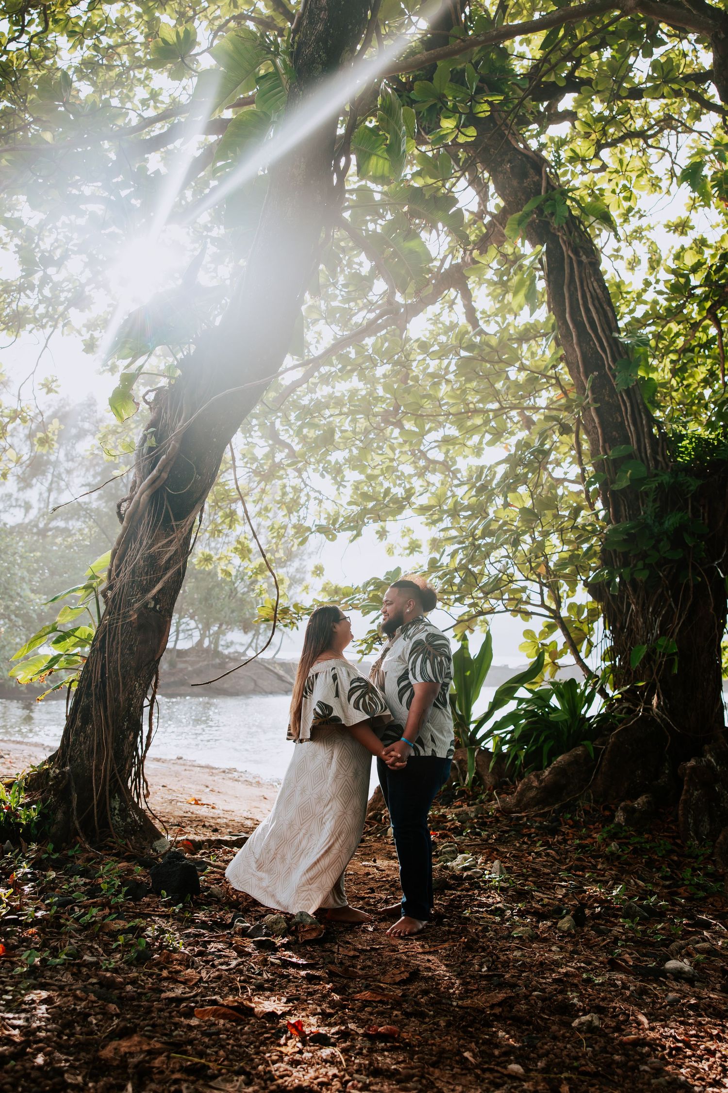 Couple holding hands under sunlit tropical trees near a beach, dressed in white and floral attire.