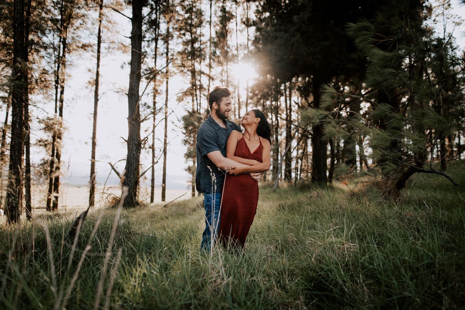 Couple embracing in a sunlit forest clearing, surrounded by tall pine trees and lush green grass.