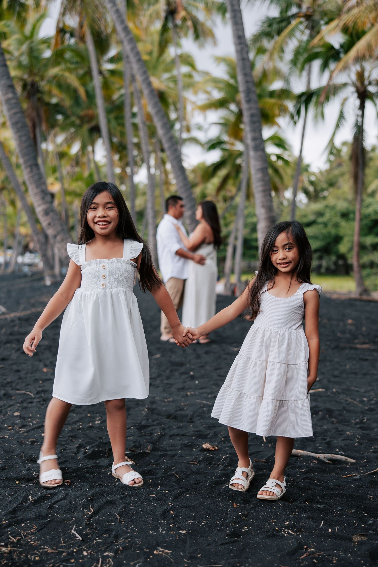 Two young girls in white dresses hold hands on black sand beach with palm trees and parents in background.