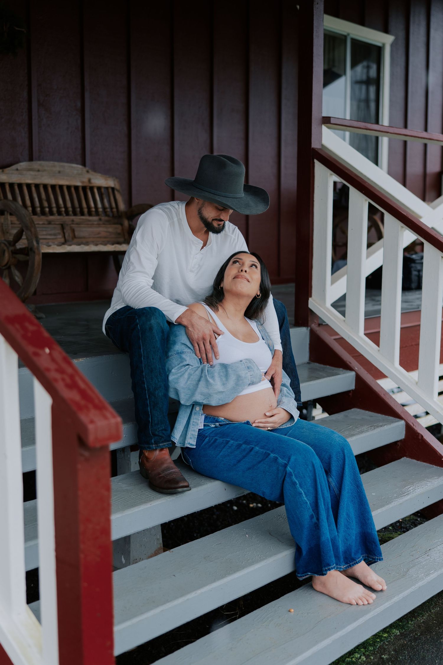 Pregnant couple sitting on porch steps, man in cowboy hat embracing woman with baby bump, rustic red farmhouse backdrop.
