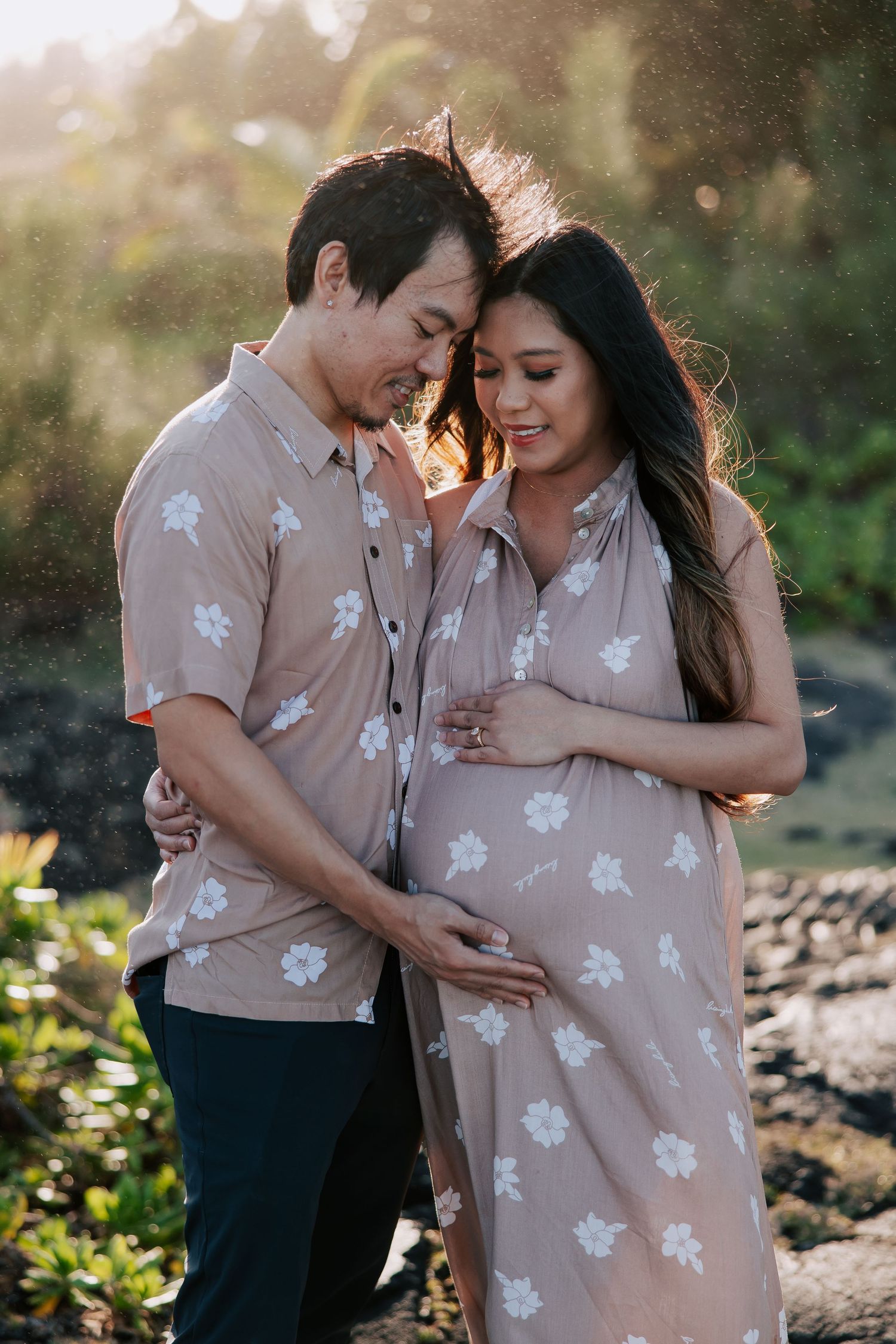 Couple in matching floral outfits sharing a tender moment outdoors, woman cradling pregnant belly at golden hour.