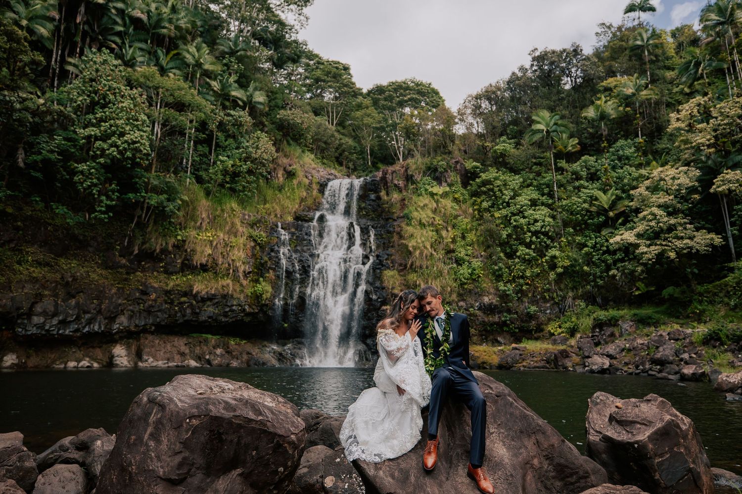 Bride and groom sitting on rocks in front of a stunning tropical waterfall surrounded by lush green jungle foliage.
