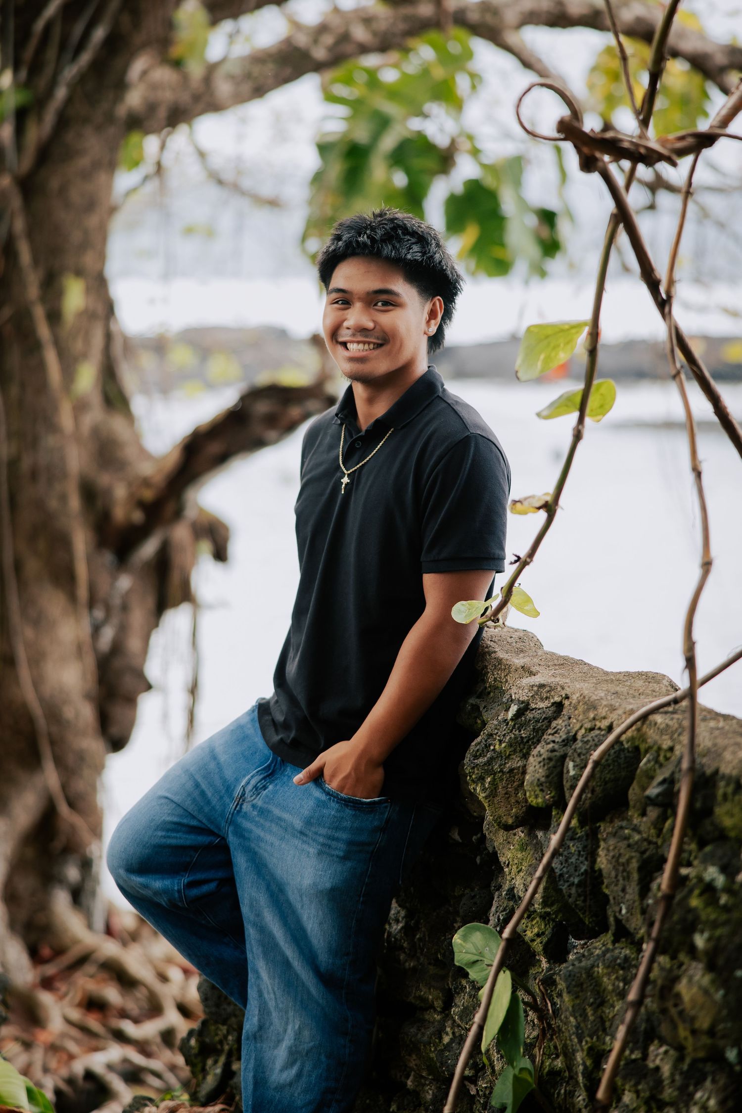 Young man in black polo and jeans smiling while leaning against a stone wall surrounded by trees near water.