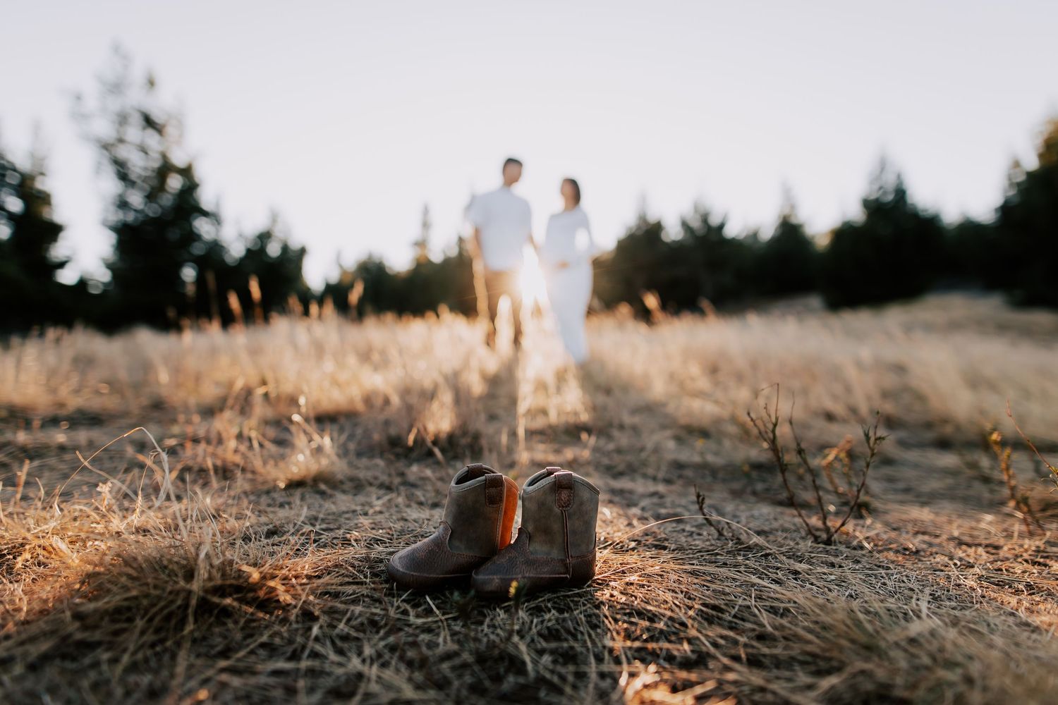 Couple stands in golden field behind tiny cowboy boots, soft bokeh sunset backdrop, rustic pregnancy announcement photo.