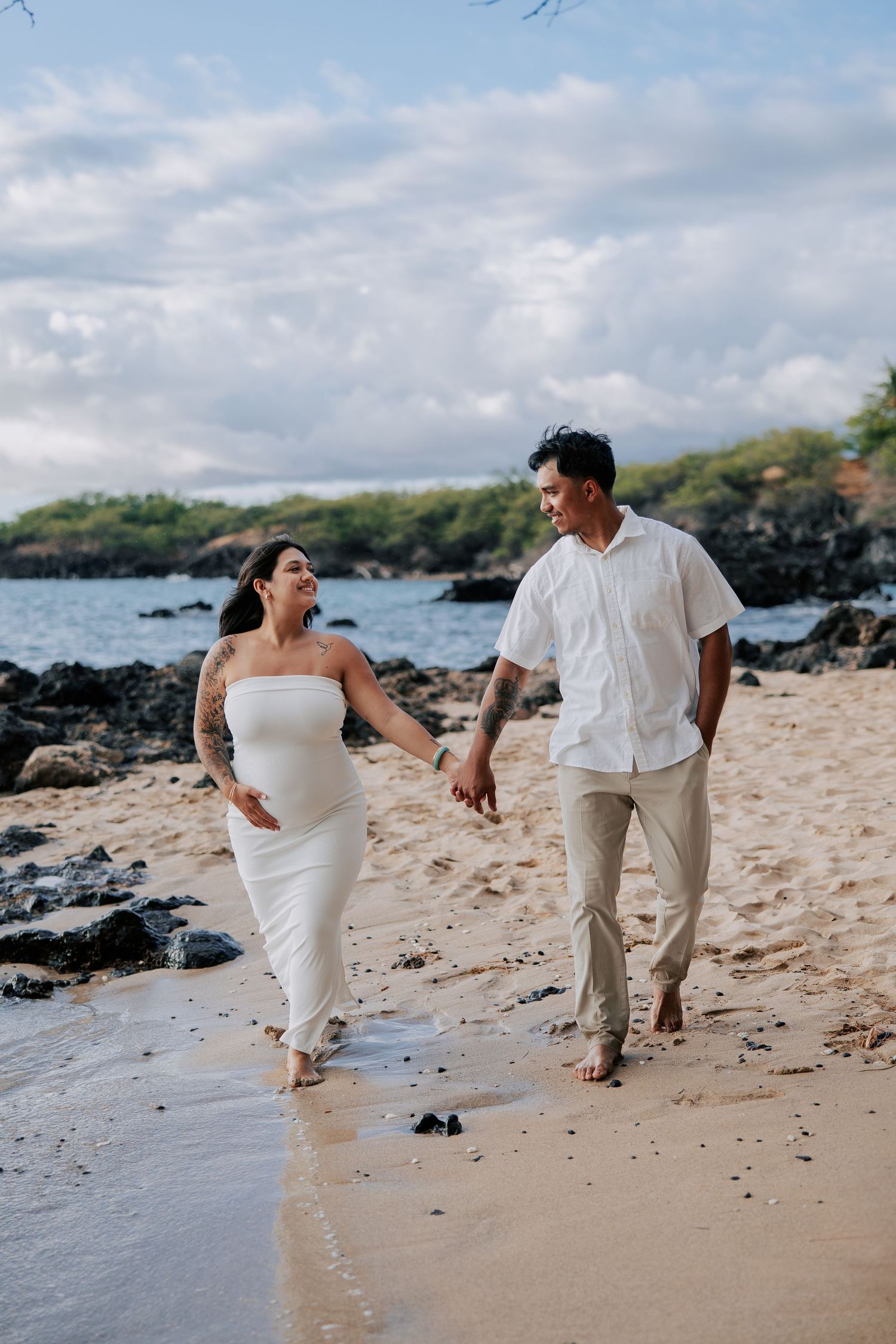 Pregnant couple walking hand-in-hand on a sandy beach, woman in white dress, man in linen shirt, rocky shoreline backdrop.