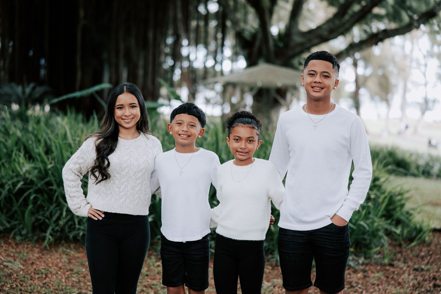 Family of four in matching white tops and black bottoms posing outdoors among lush green trees.