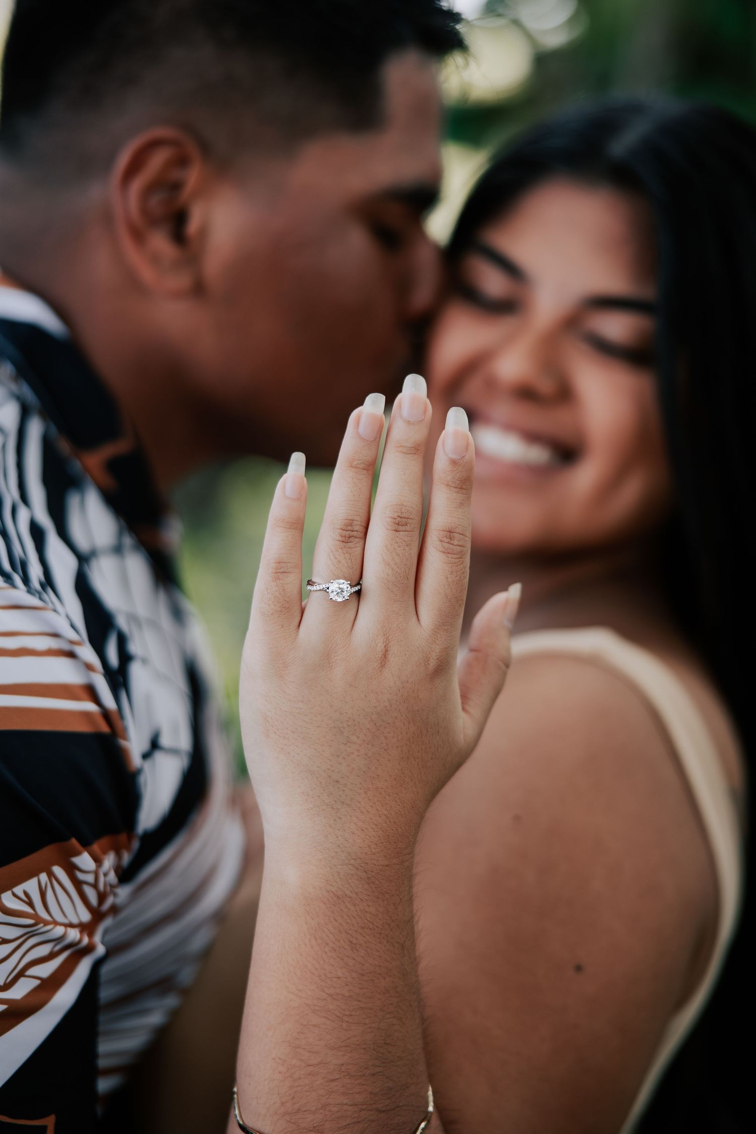 Couple celebrating engagement with woman smiling and showing diamond ring while man kisses her cheek.