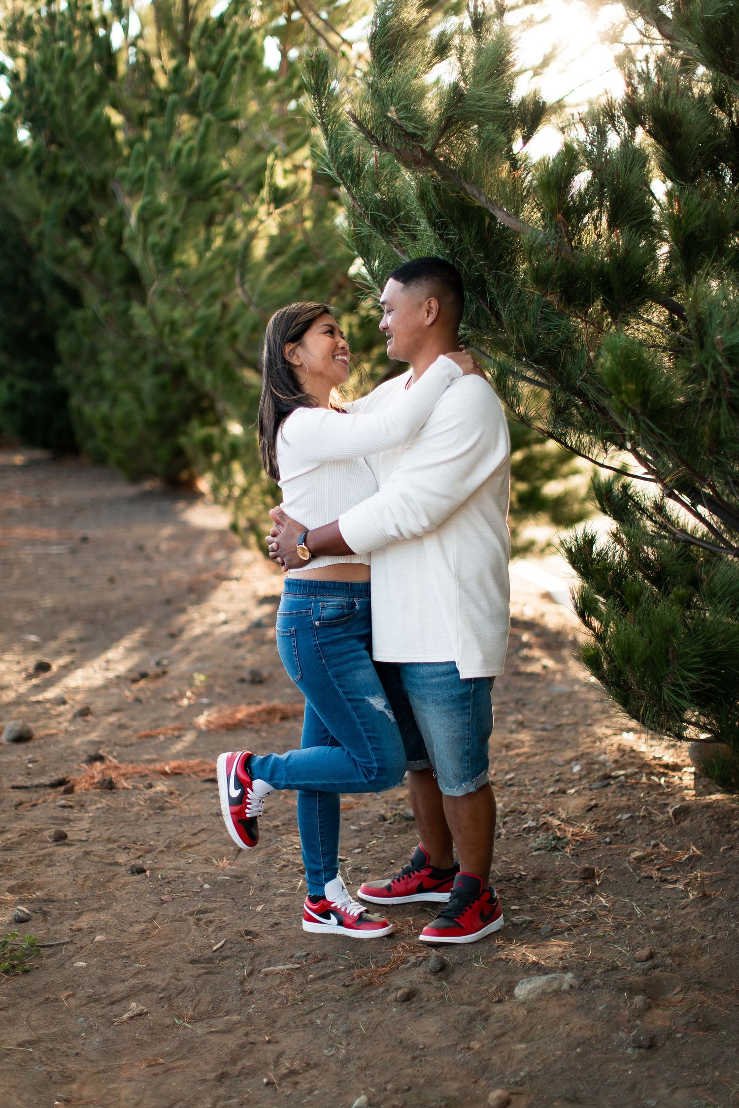 A happy couple embraces and smiles at each other among pine trees, both wearing white tops, jeans, and red sneakers.