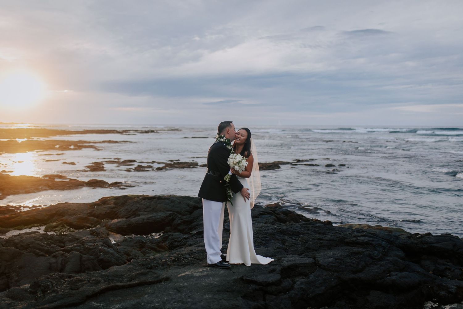 Couple sharing a romantic kiss on rocky coastal shore at sunset, bride in white gown holding bouquet, groom in dark suit.